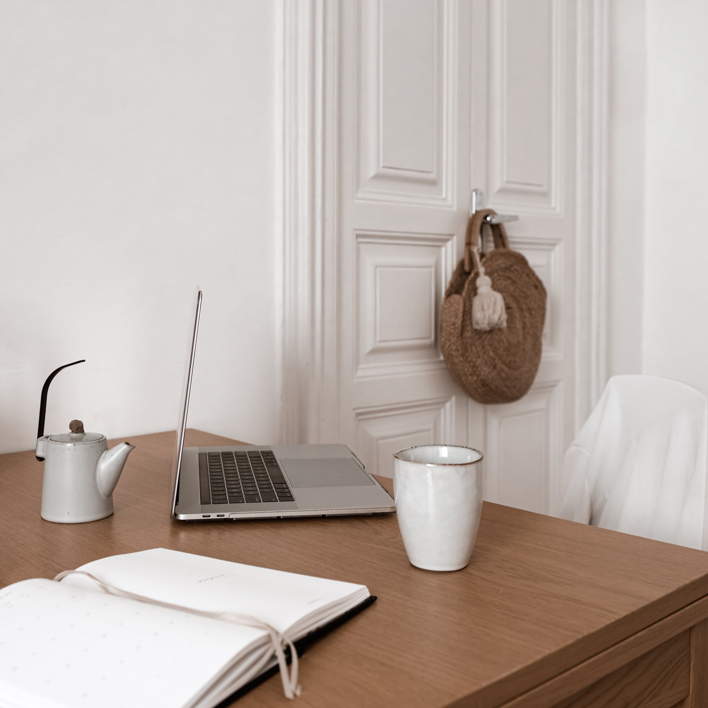 A wooden table holding a white teapot, an open notebook, a laptop, and a white mug with a gold rim, with white walls and a closed white door in the background.