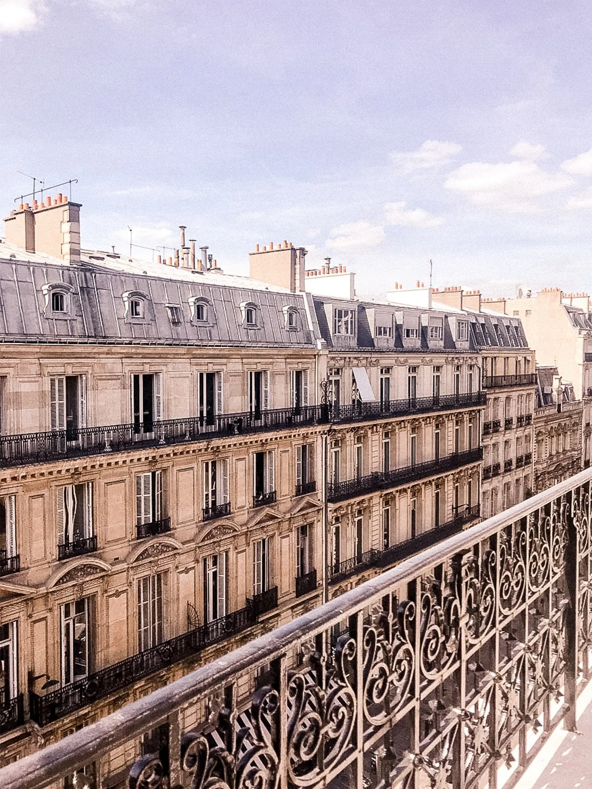 View from a balcony showing classic Parisian apartment buildings with ornate facades, tall windows, and wrought iron balconies under a cloudy sky.