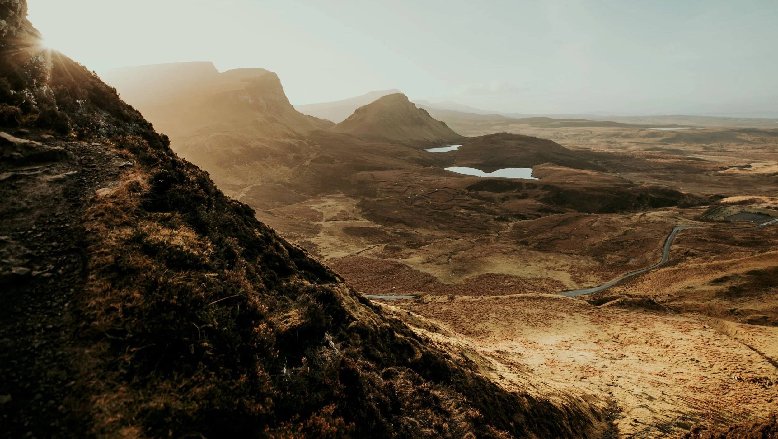 A mountainous landscape with steep hills, two small lakes, and a winding road under a cloudy sky with the sun shining from behind the mountains.