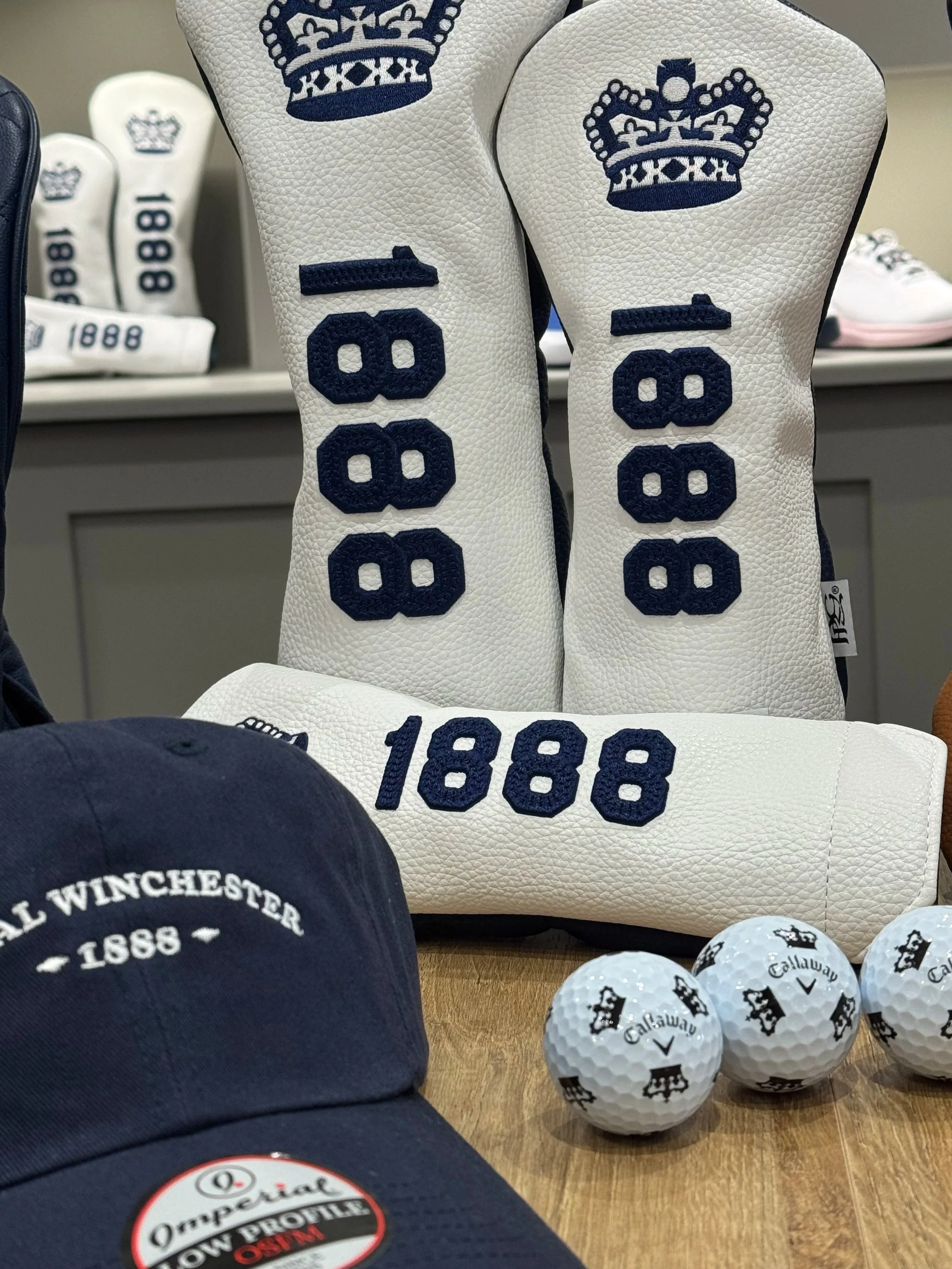 Golf accessories including white golf headcovers with navy crown and '1888' embroidered, a navy cap with white 'Royal Winchester 1888' embroidery, several white Callaway golf balls, and a few other golf accessories on a wooden surface.