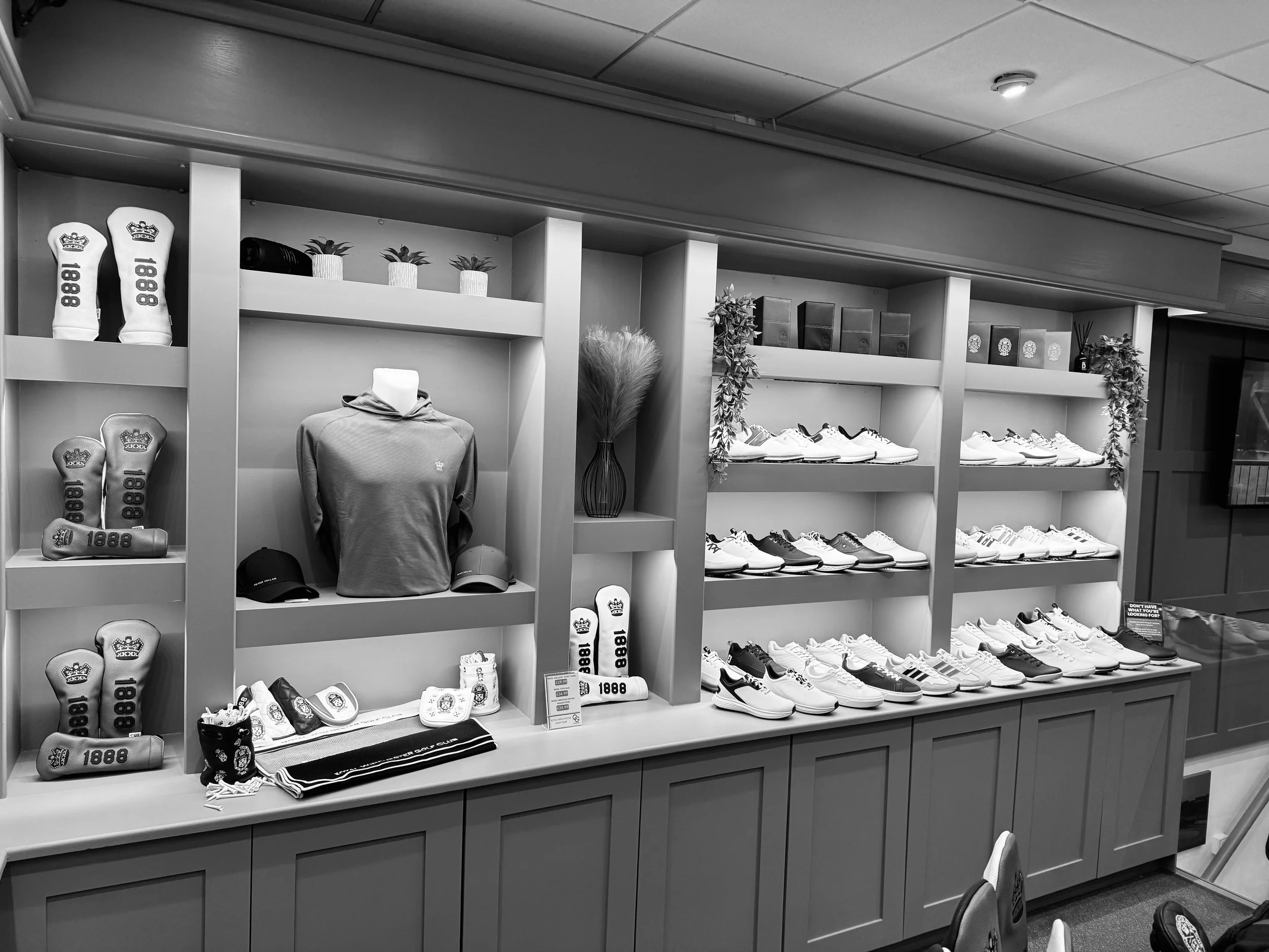A display shelf in a retail store featuring golf shoes, golf balls, and accessories. The shelf has multiple rows, with shoes on one side and accessories like hats and towels on the other. There are decorative plants and boxes on the top shelf.