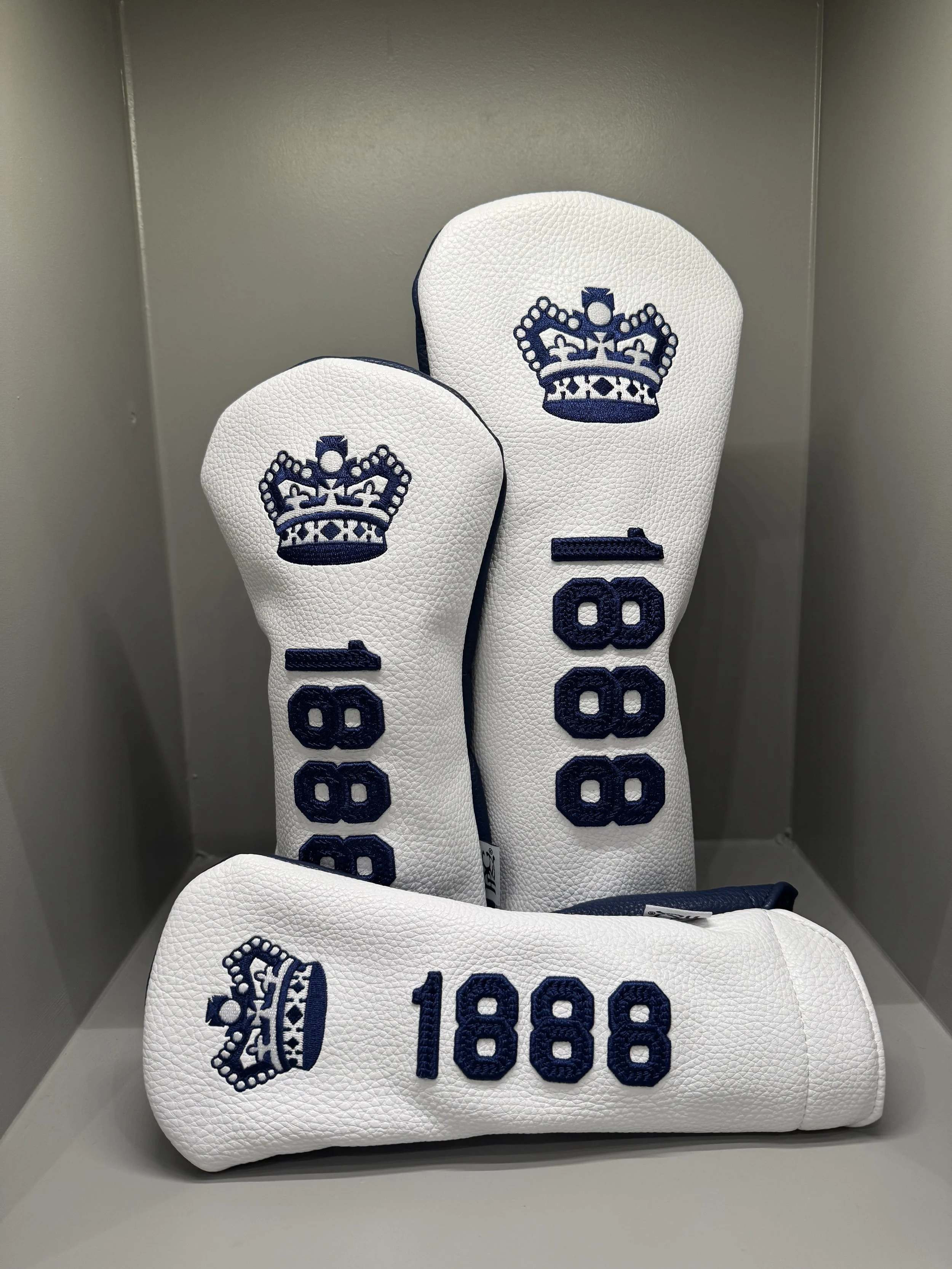White golf club headcovers with navy blue embroidered crown and the numbers 100 and 1888, placed inside a gray metal cabinet.