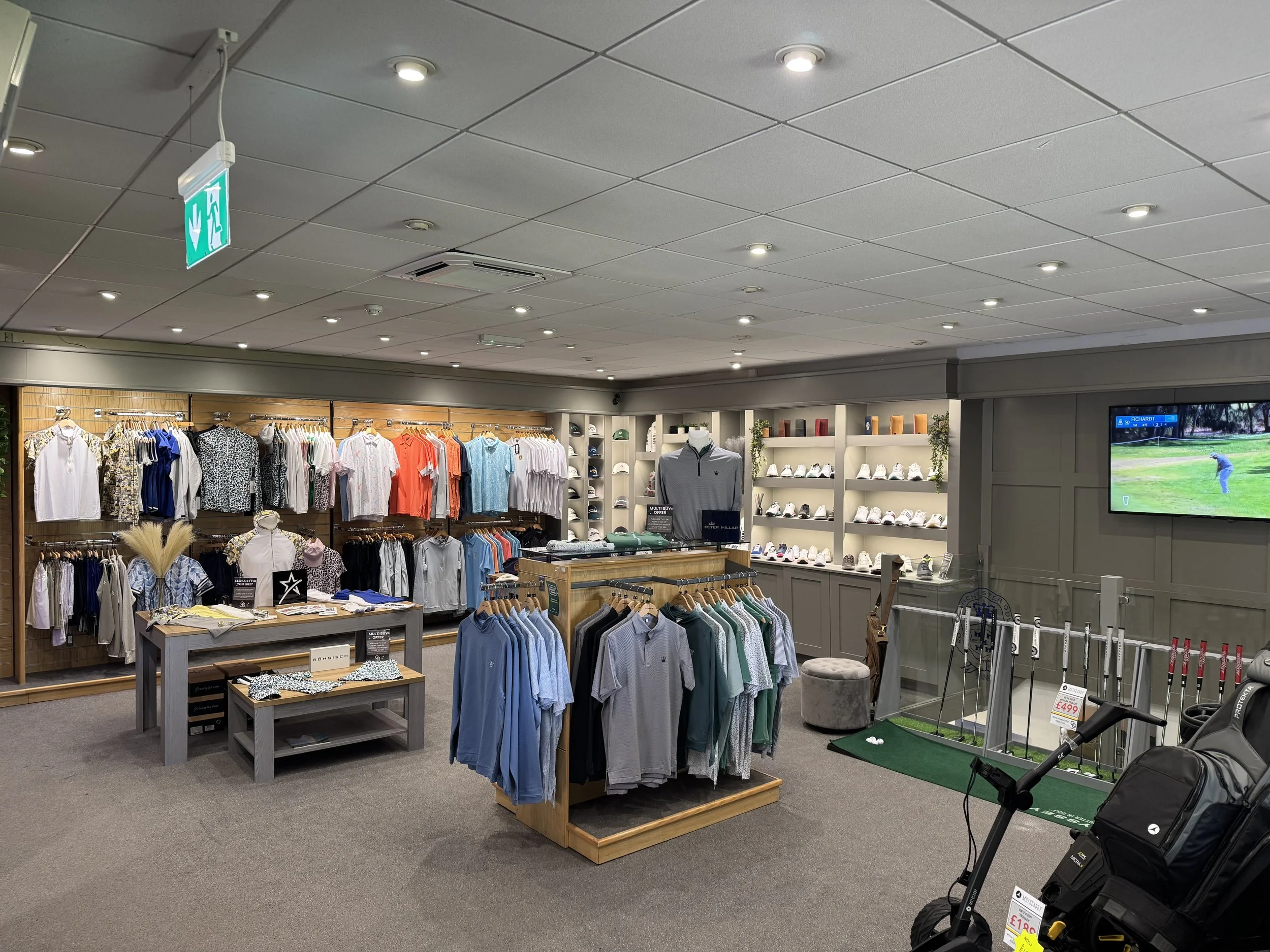 Inside a retail store displaying men's casual and golf apparel, shoes, and golf clubs, with signage and a television showing a golf game.