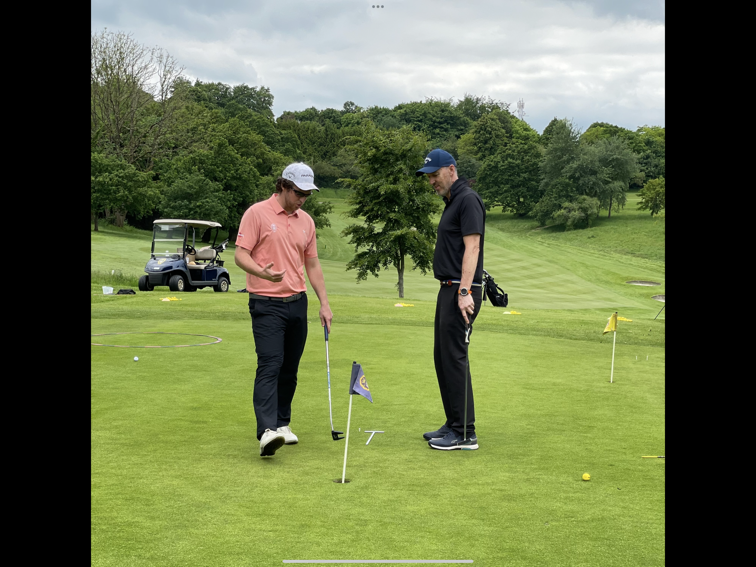Two men are on a golf course discussing putting. One is in a pink polo shirt and cap, holding a golf club, and the other is in a black shirt, holding a golf club. There is a golf cart in the background, along with green trees and a cloudy sky.