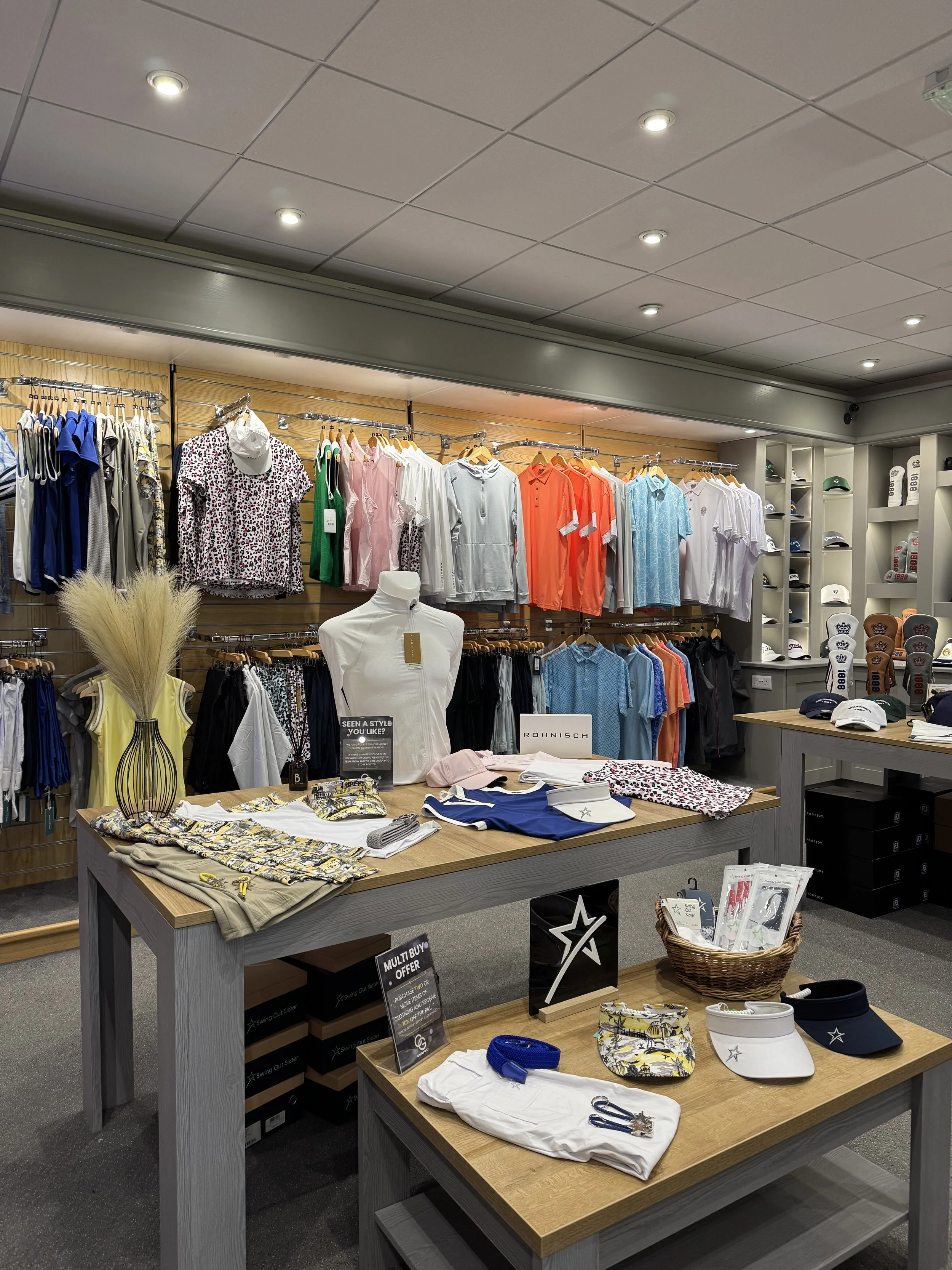 Inside a clothing store, display tables and racks with various men's and women's apparel, including polo shirts, hats, and accessories.