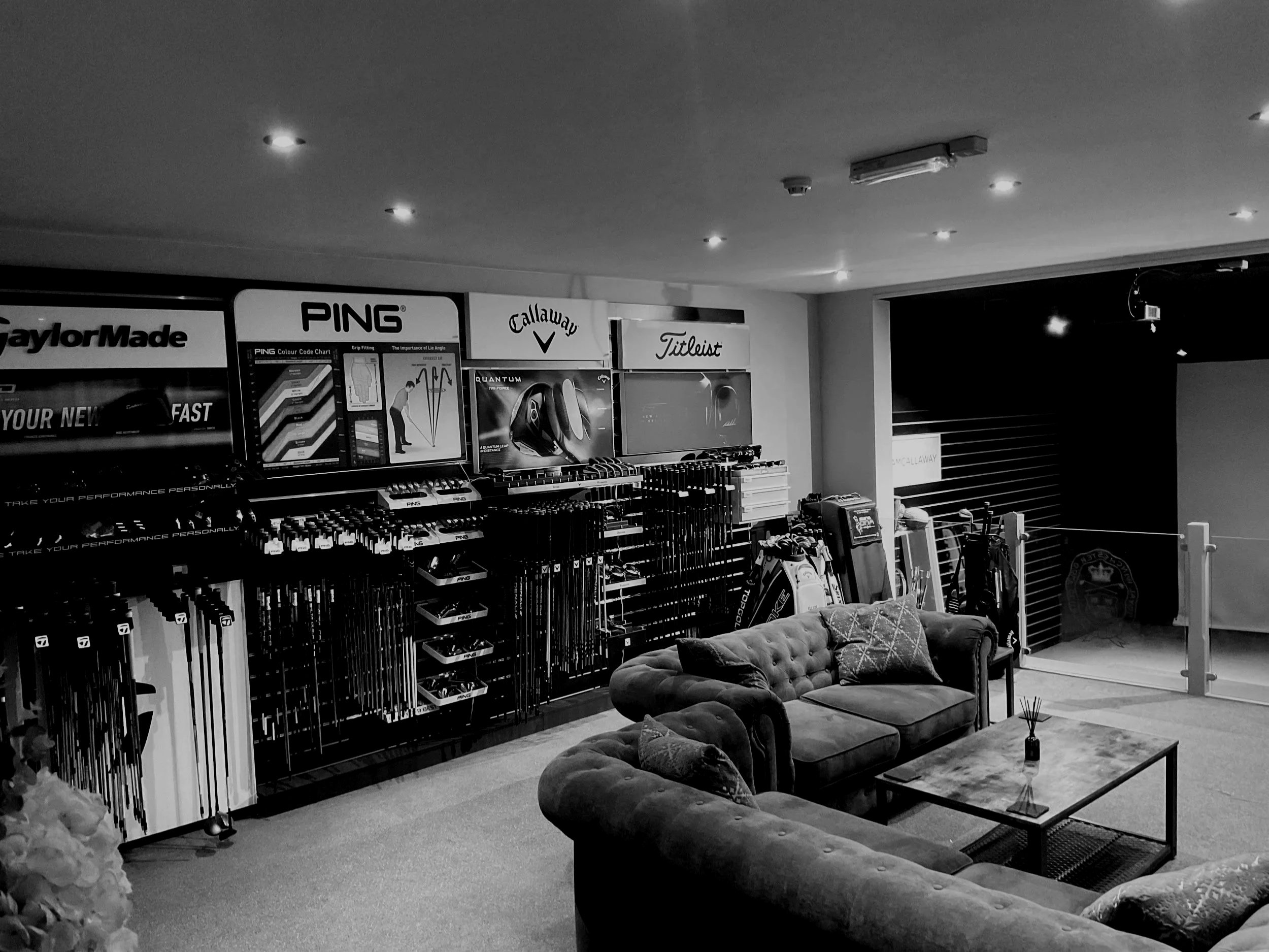 A retail showroom with golf equipment and accessories on display, including golf clubs, golf balls, and brands like Ping, Callaway, TaylorMade, and Titleist. There is a lounging area with velvet tufted sofas and a coffee table with a small decorative plant.