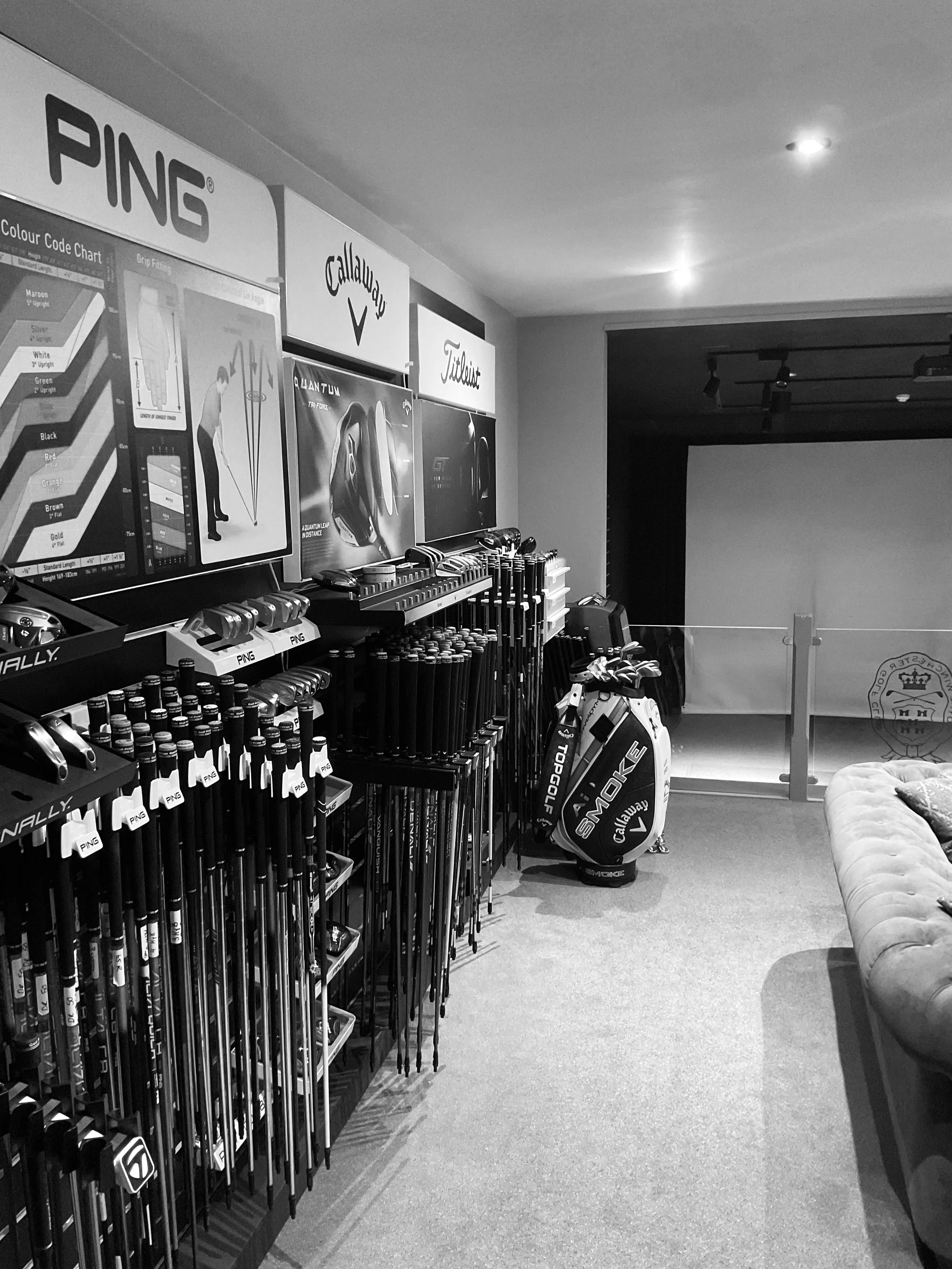 A golf retail store display with clubs, golf bags, and signage for brands like Ping, Callaway, and Titleist, in a room with a couch and a projection screen.