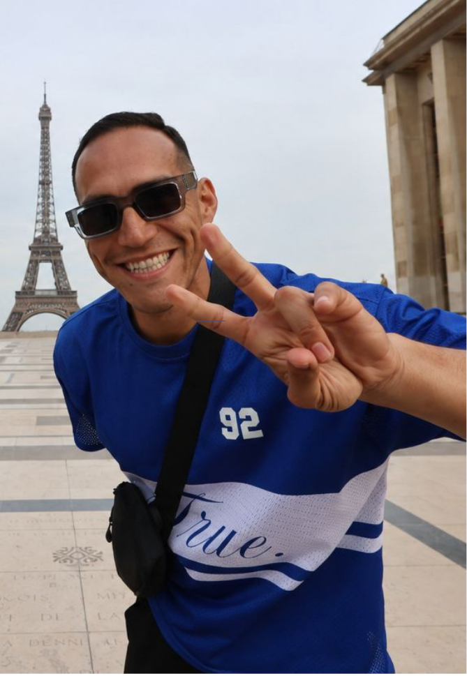 Un hombre sonriente con gafas de sol haciendo el signo de la paz con los dedos frente a la Torre Eiffel, en París, Francia.