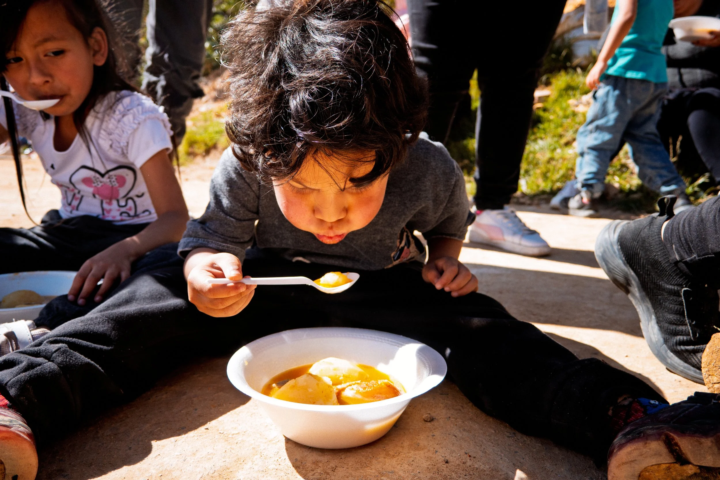 Unos niños sentados en el suelo al aire libre, compartiendo una comida de un cuenco con una cuchara. La imagen se centra en un niño de pelo oscuro y rizado que come con la cuchara, rodeado de otros niños y adultos.