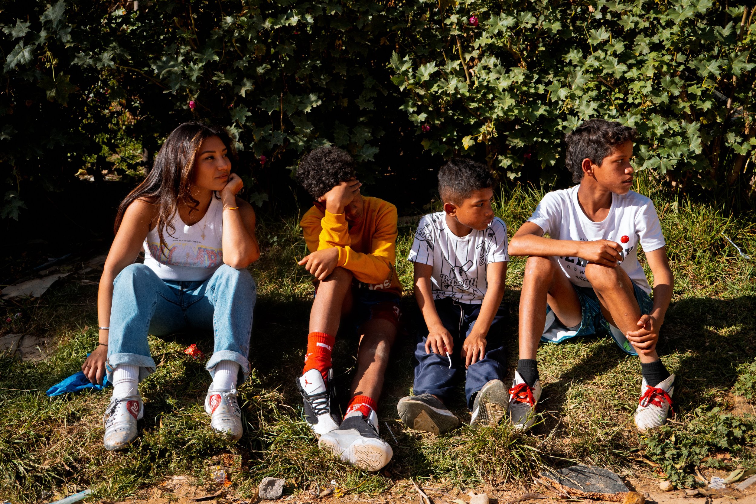 Ivana Rojas y tres niños sentados al aire libre frente a unos arbustos de hojas verdes, con la luz del sol iluminándolos.