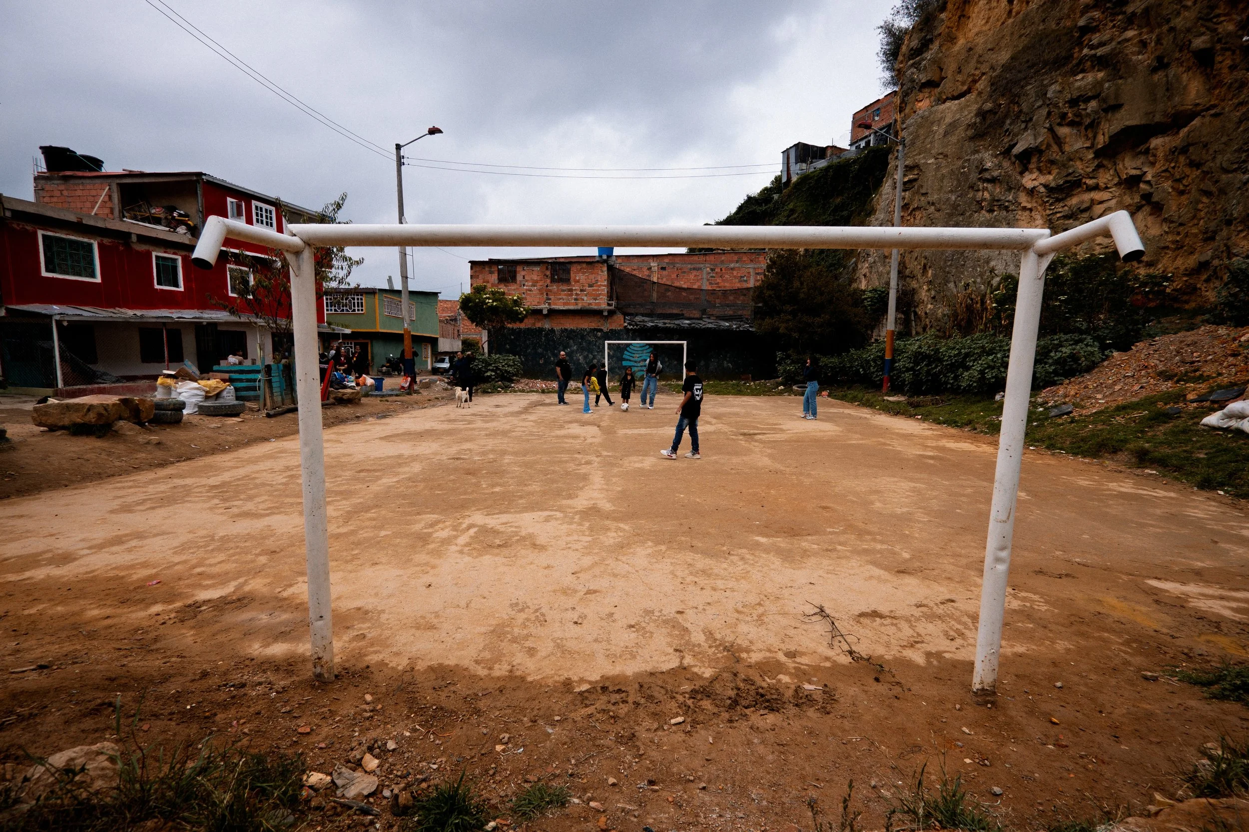 Niños y adultos jugando al fútbol en un campo de tierra, en un barrio residencial con casas de colores y una ladera rocosa al fondo.