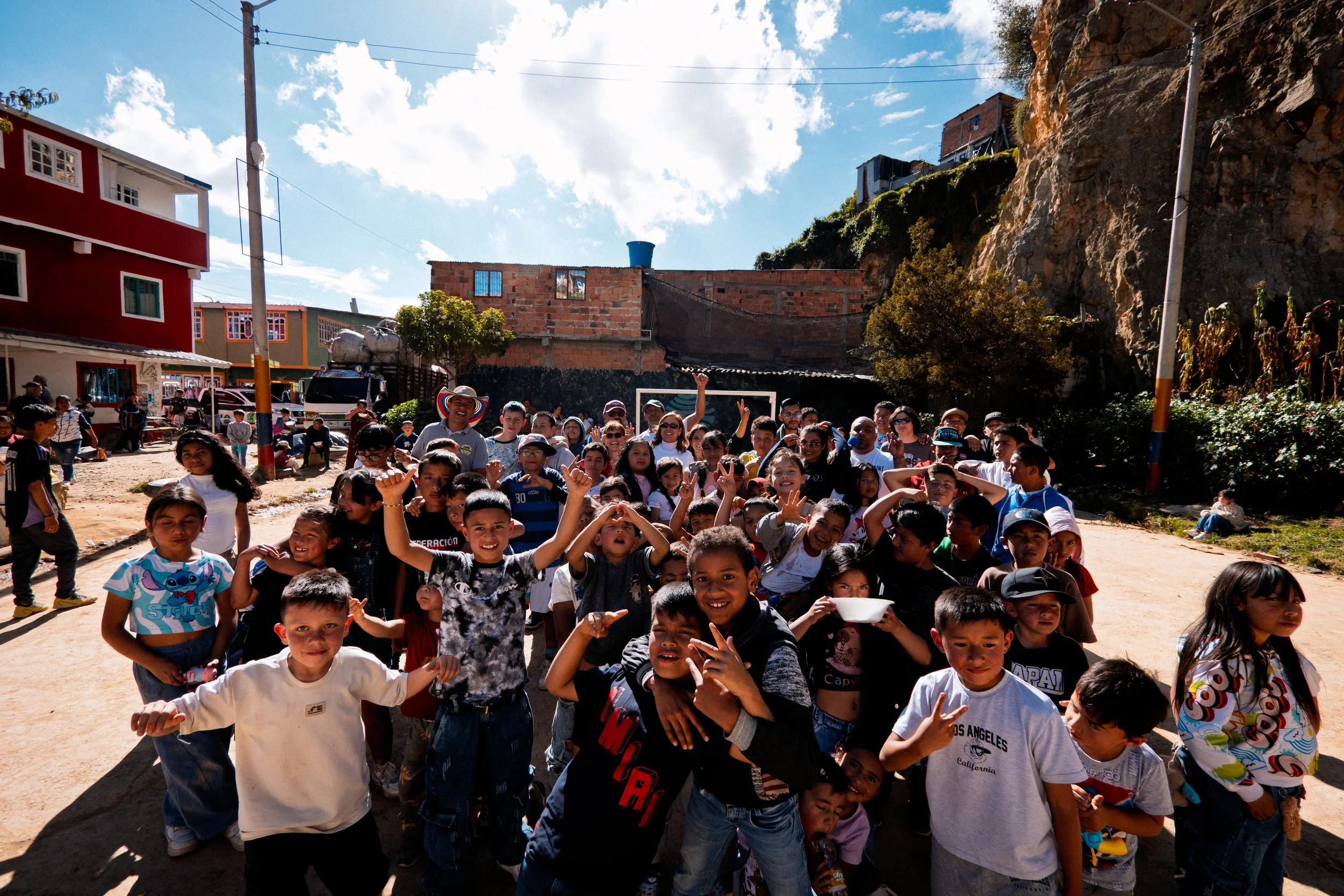 Un grupo de niños y adultos se reunía al aire libre en una zona comunitaria, sonriendo y haciendo el signo de la paz, con edificios de colores y un paisaje montañoso al fondo.