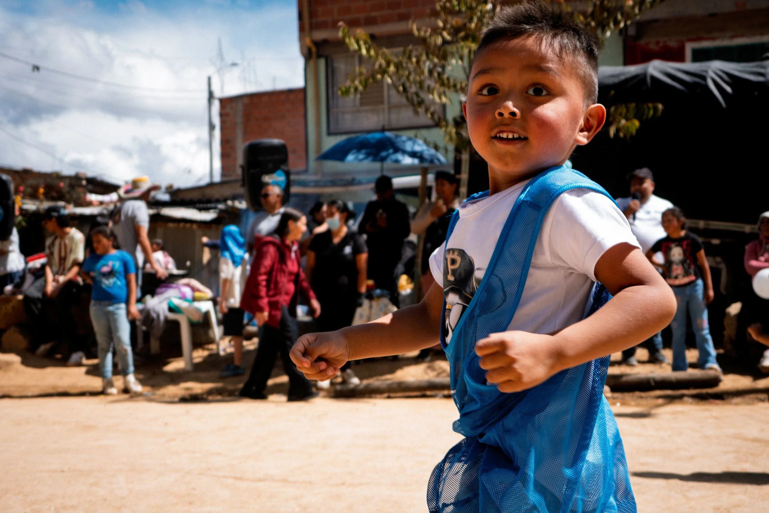 Un niño pequeño con una camiseta blanca y una mochila azul que corre al aire libre, con gente paseando y charlando al fondo.
