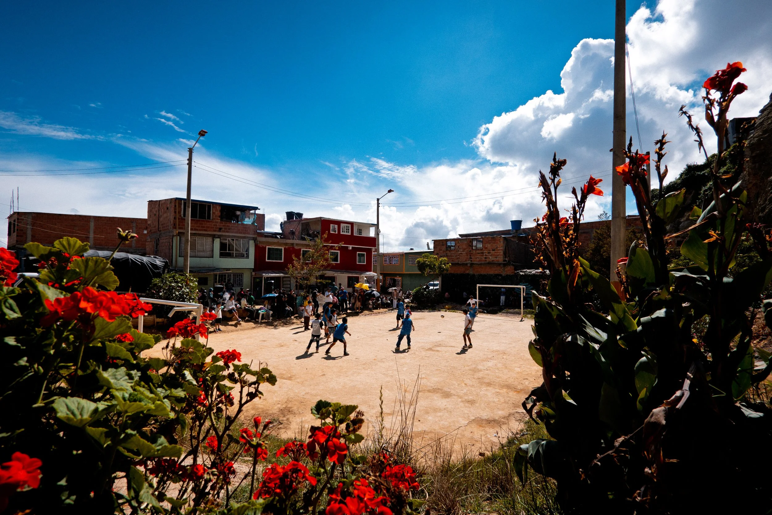 Niños jugando al fútbol en un campo de tierra en un barrio con casas, algunas de ellas en construcción, bajo un cielo parcialmente nublado.