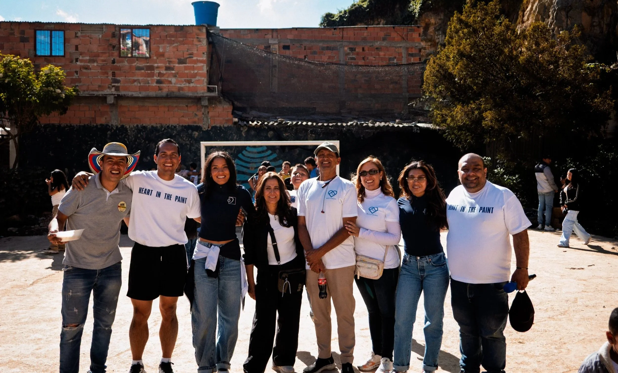 Un grupo de ocho personas de diferentes orígenes, de pie al aire libre, sonriendo, con un edificio de ladrillo, árboles y otras personas al fondo.