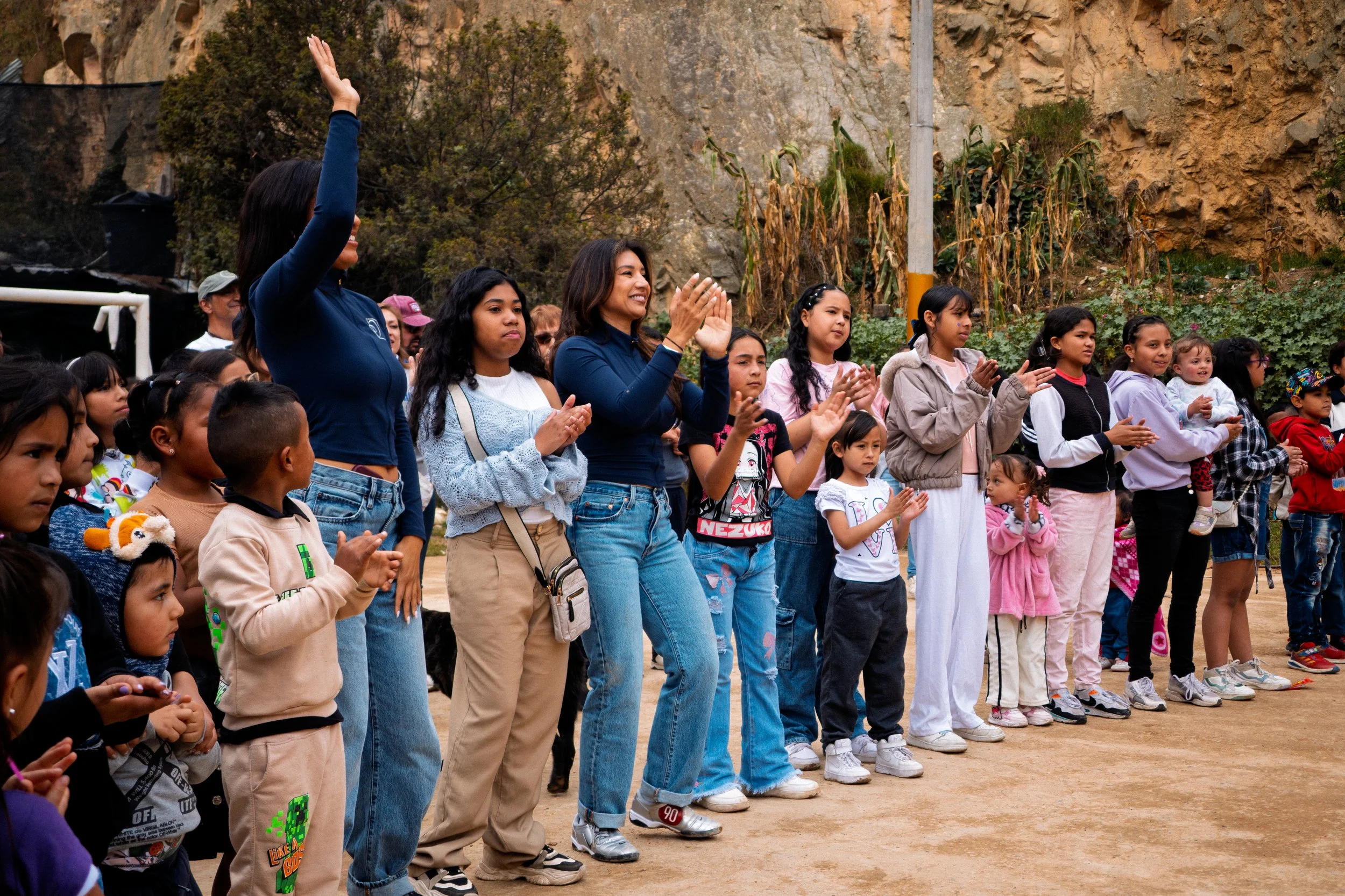 Un grupo de niños y dos mujeres adultas de pie al aire libre, aplaudiendo y levantando las manos, con un acantilado rocoso y plantas de maíz secas al fondo.