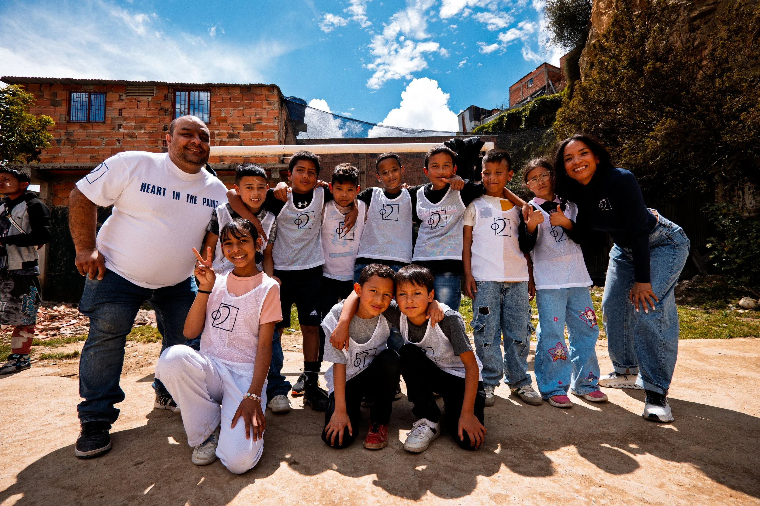 Un grupo de niños y dos adultos posan al aire libre delante de unas casas; algunos niños llevan camisetas deportivas blancas con números, bajo un cielo parcialmente nublado.