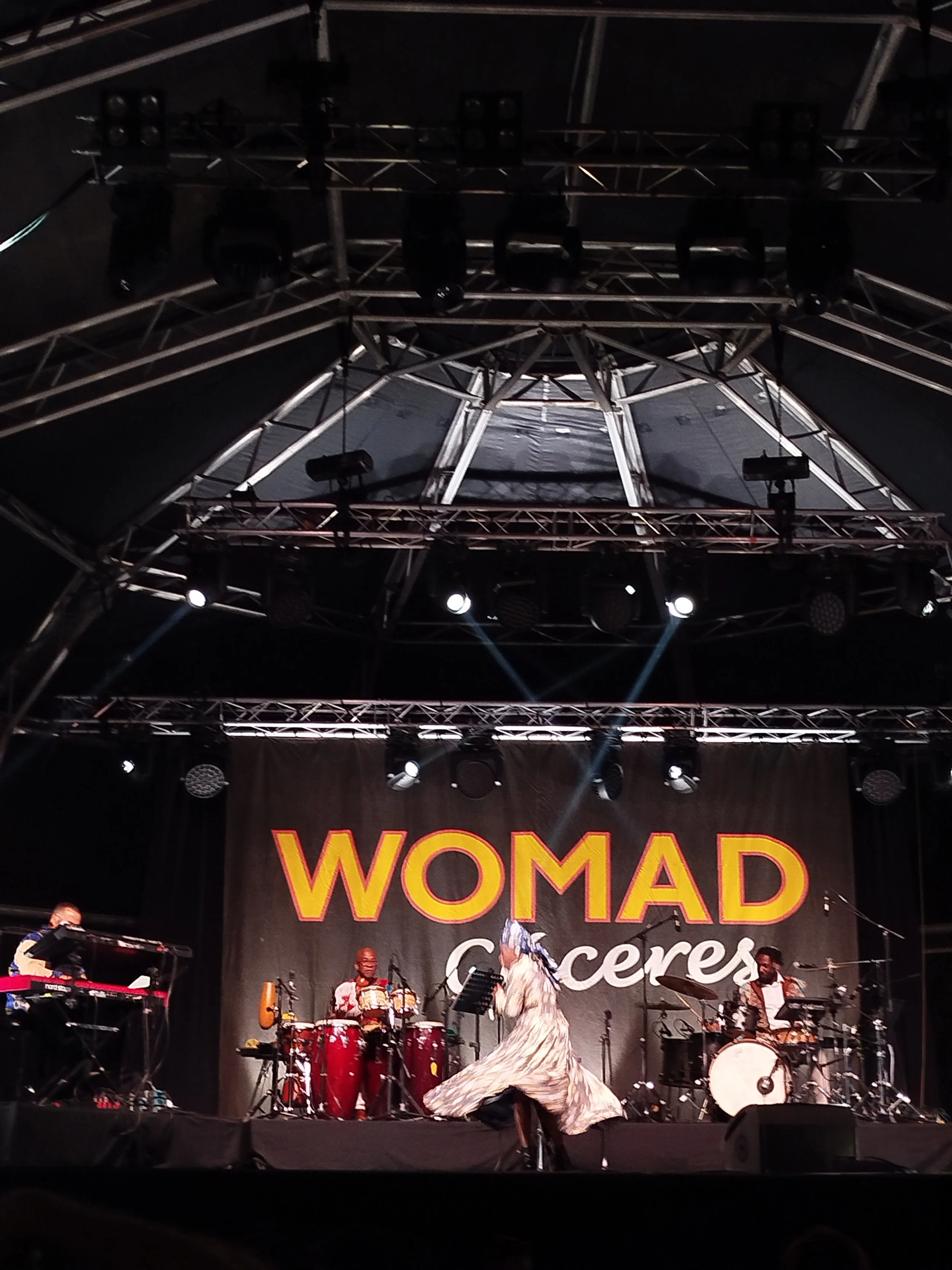 Musicians performing on stage at a Women conference, with instruments including drums, keyboard, and microphones, under stage lighting and a large banner that reads 'WOMEN'.