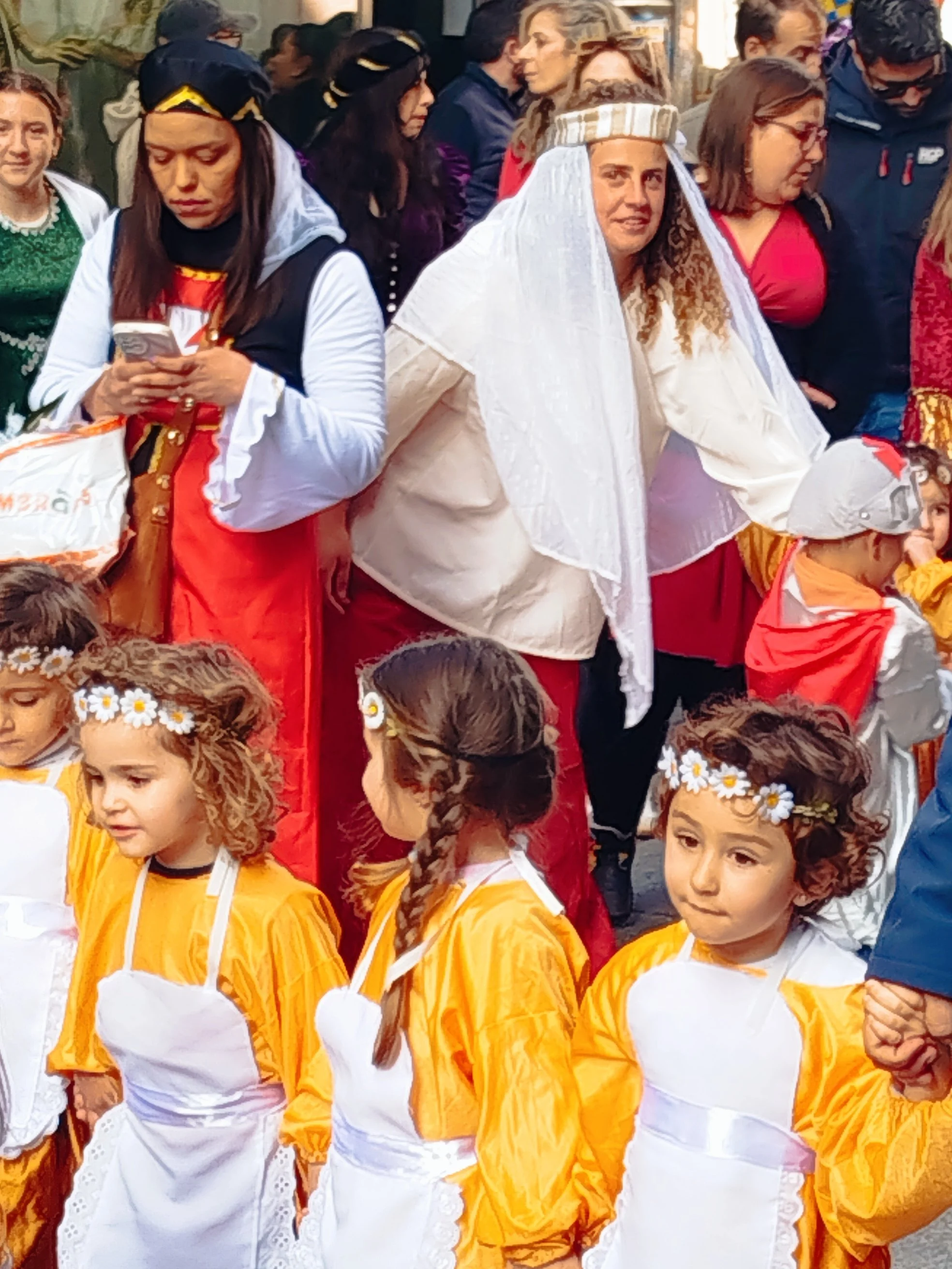 Group of children and adults dressed in colorful costumes, with some women wearing traditional or festive attire, gathered in a crowd for a celebration or parade.