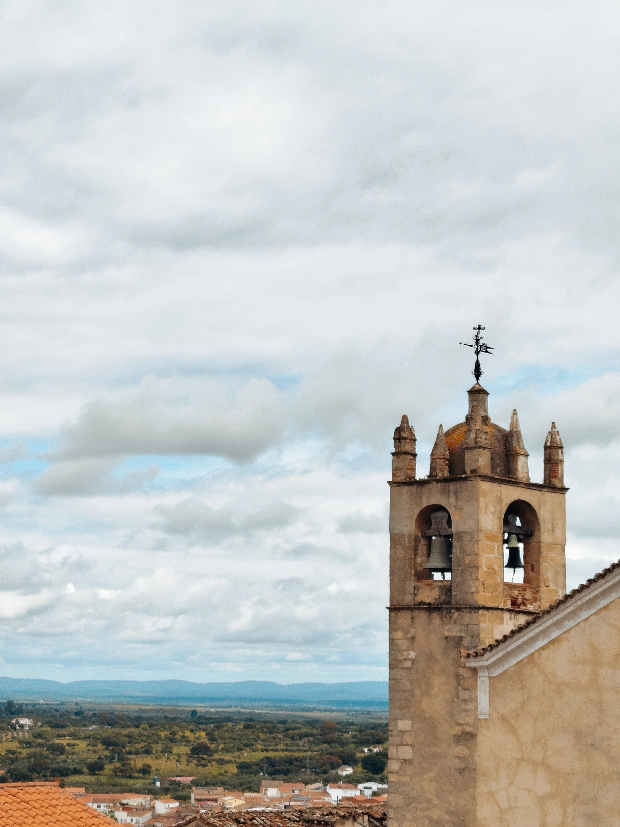 A historic church with a bell tower, topped with a weathervane, overlooking a rural landscape under a cloudy sky.
