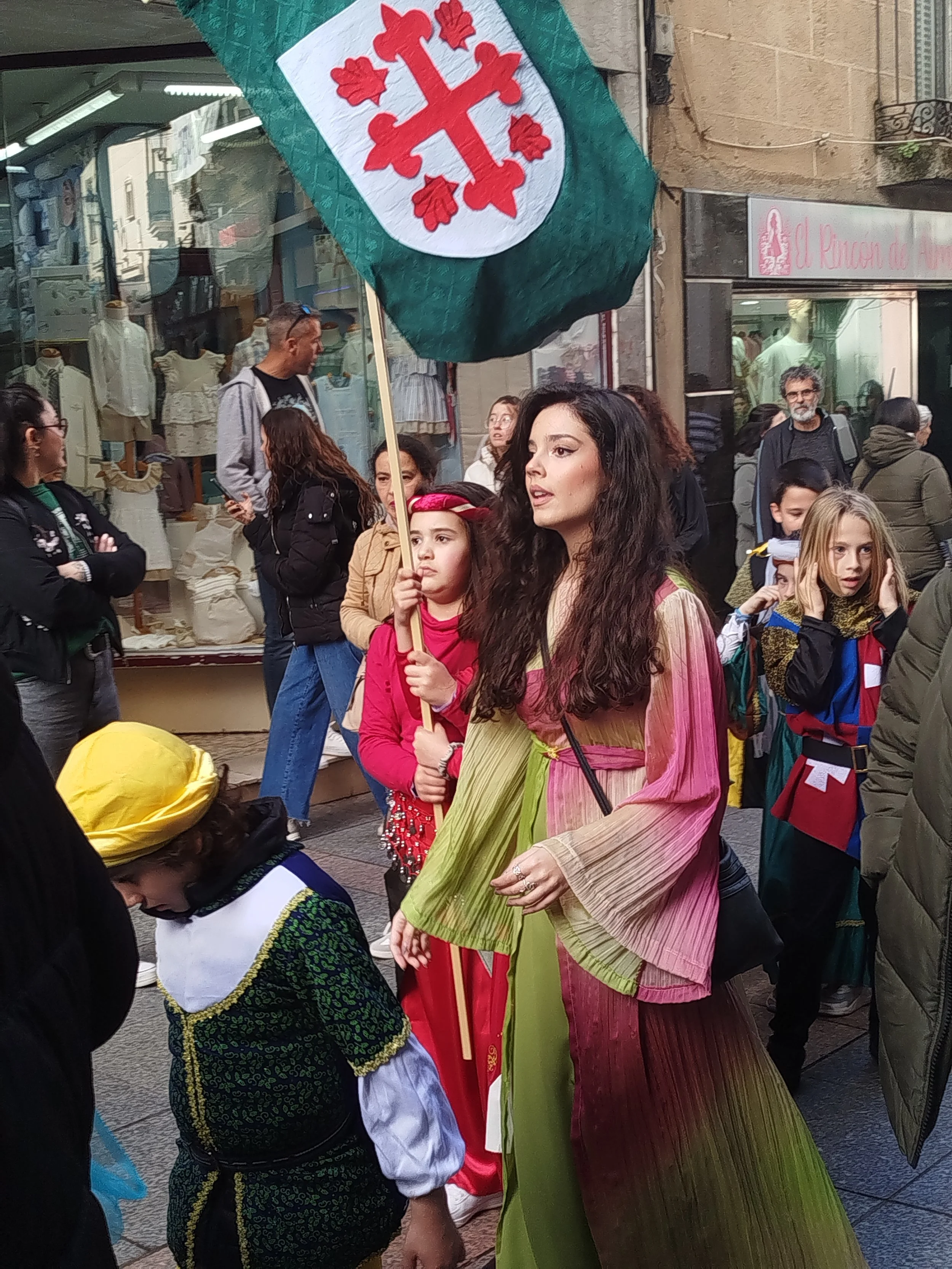 Street scene with people, a woman wearing a pink and green dress holding a flag with a red cross and four red symbols, and a young girl in traditional attire. Background includes shopfronts and passersby.