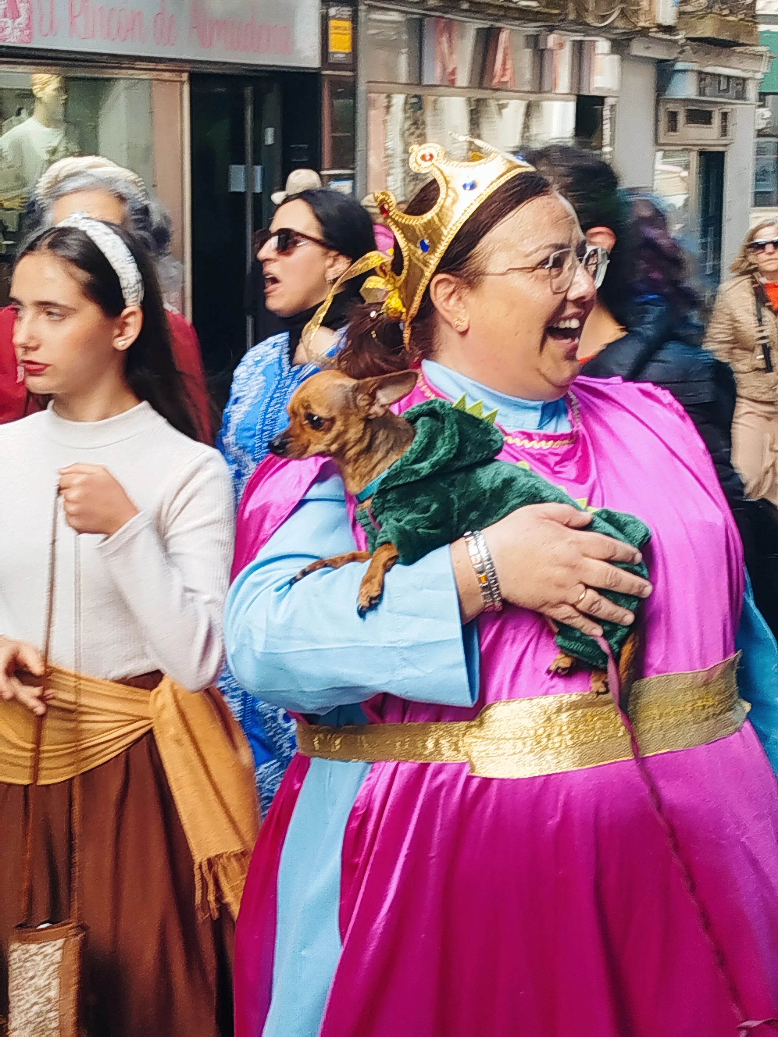 A woman in a pink and light blue dress, wearing a golden crown and glasses, holding a small dog dressed in a green outfit. She is smiling joyfully in a crowded street scene.