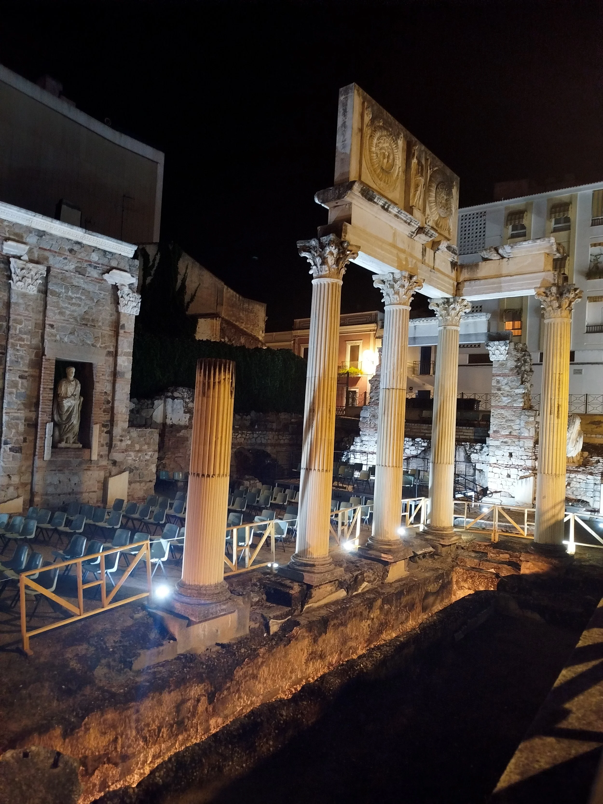 Nighttime view of ancient Roman ruins with four fluted columns and intricately carved architrave, illuminated by artificial lights, with modern buildings in the background.