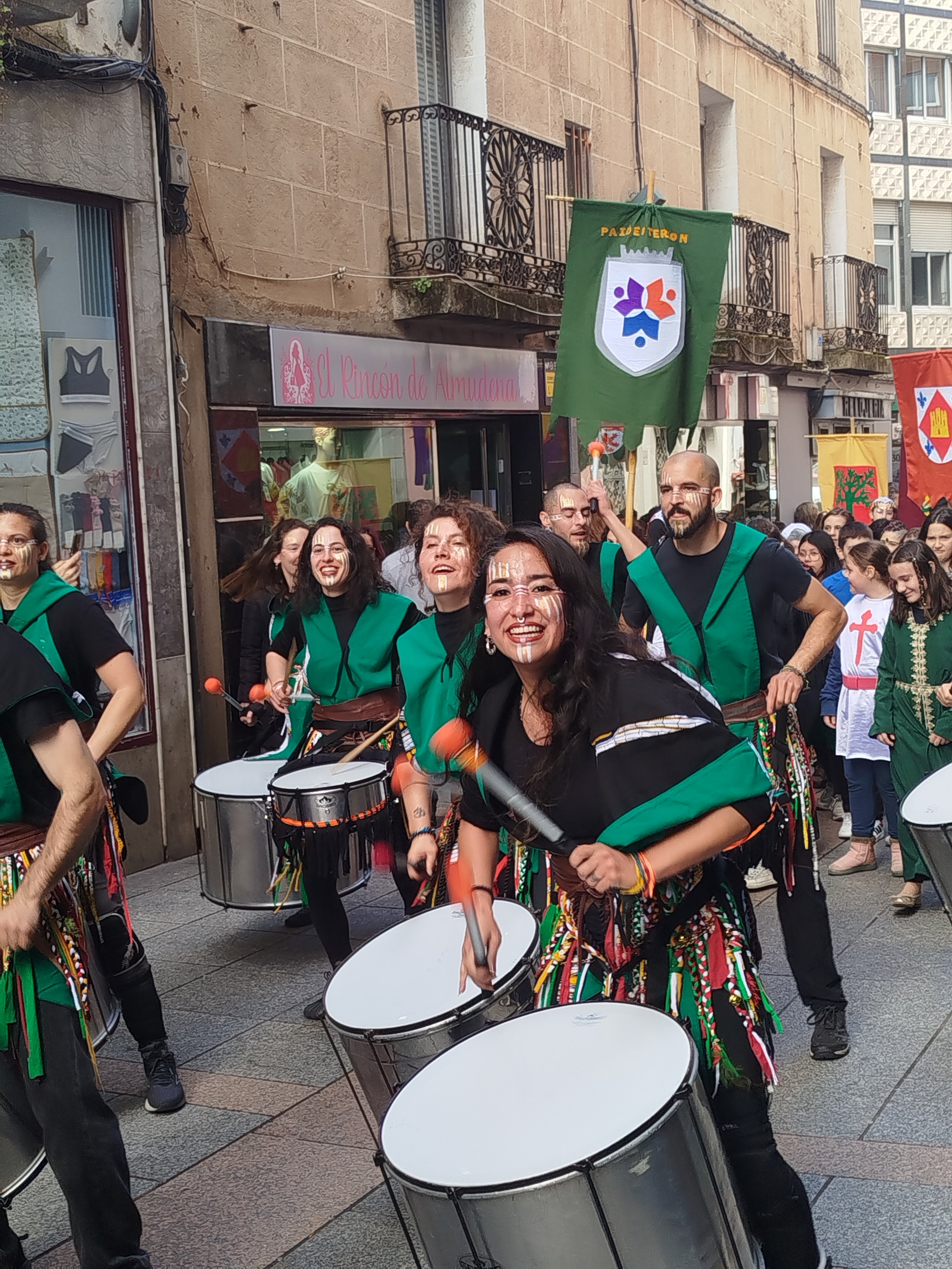 A diverse group of people participating in a parade or festival, dressed in colorful traditional clothing with face paint, playing drums and other percussion instruments on a city street with buildings and balconies in the background.