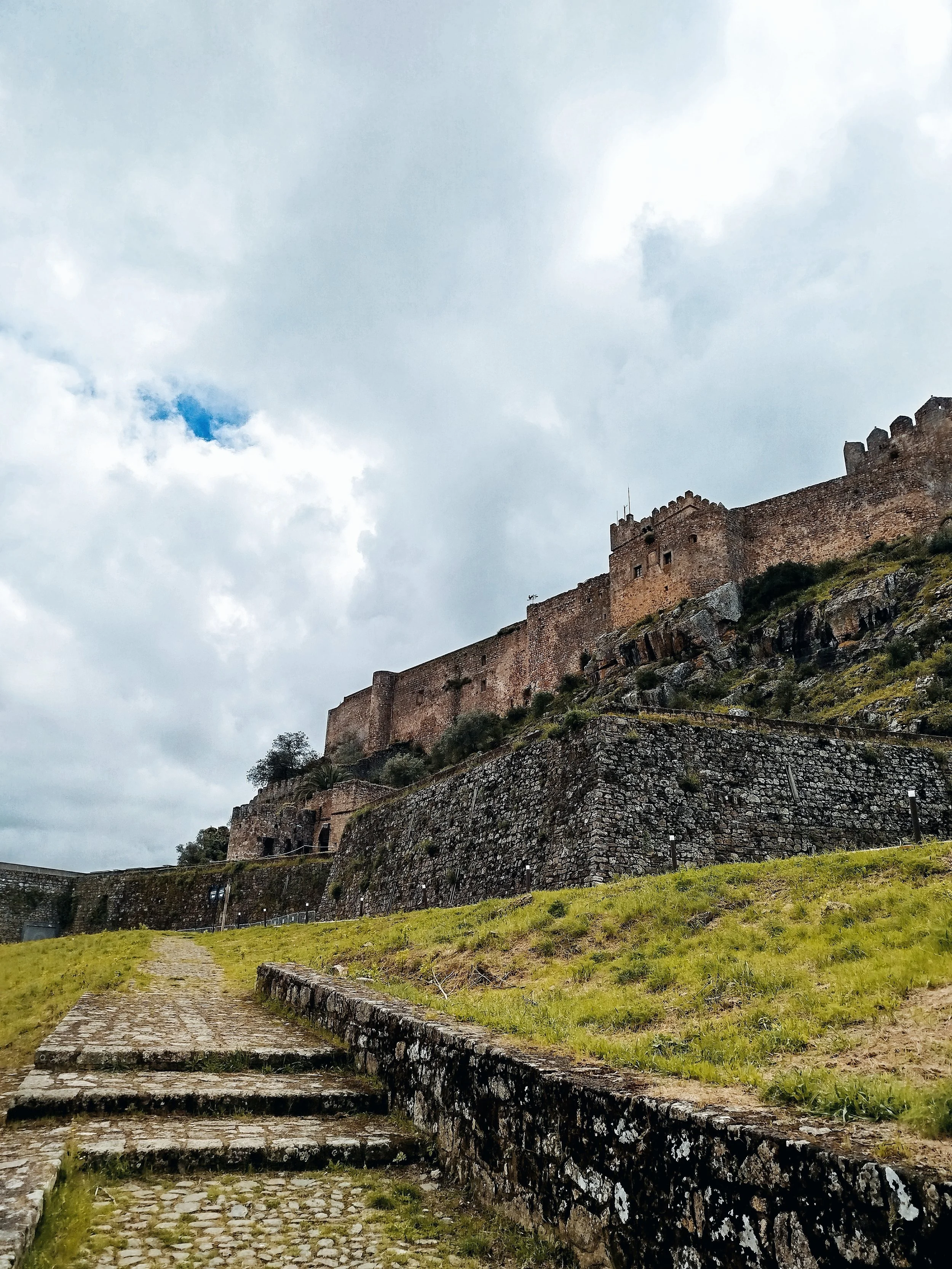 Ancient stone castle on a hill overlooking a cobblestone pathway and grassy area, cloudy sky overhead.