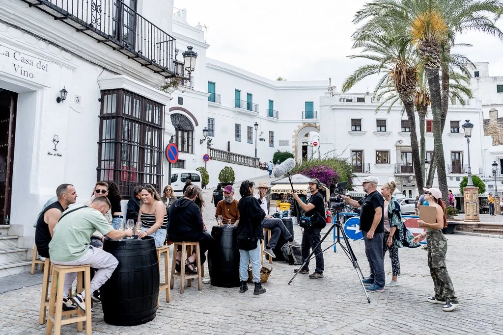 People filming a video or interview outside on a street, with some sitting on barrel stools around a table, and others standing, with a camera operator recording. White buildings, palm trees, and a cloudy sky in the background.