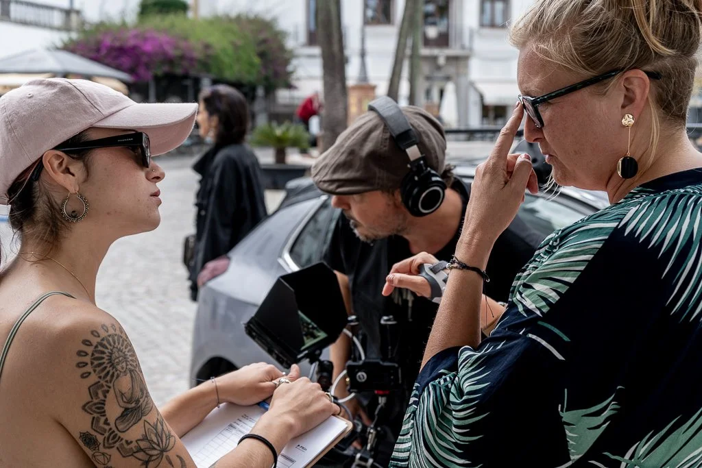 A woman with tattoos and sunglasses is talking to another woman with glasses, wearing a patterned top, while a man with headphones and a camera is taking photographs in a street with cars and pedestrians.