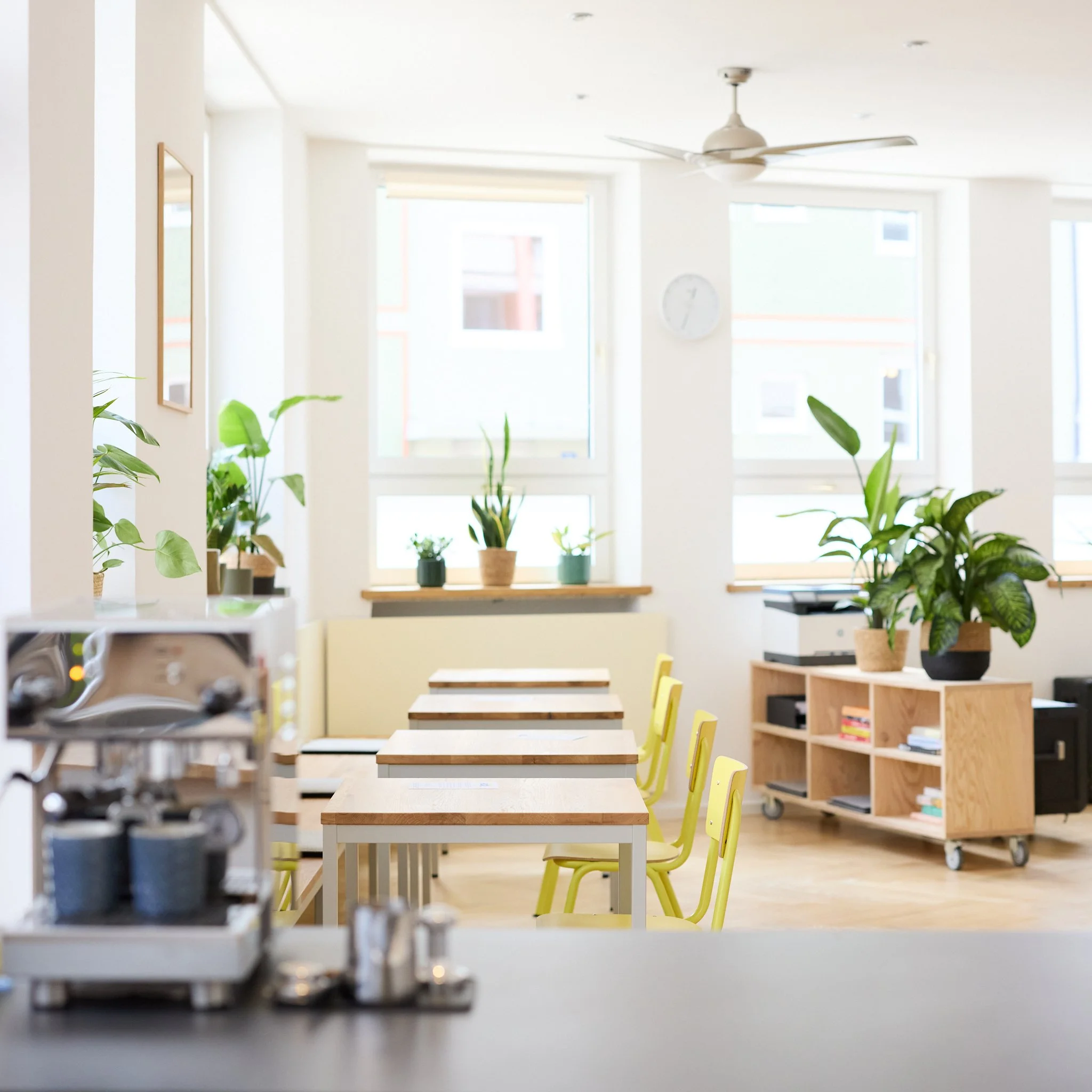 Helle, modern ausgestattete Küche oder Esszimmer mit Holz- und Kunststoffmöbeln, mehrere Tische mit gelben Stühlen, große Fenster, Pflanzen auf der Fensterbank, Uhren an der Wand, und eine Deckenventilator.