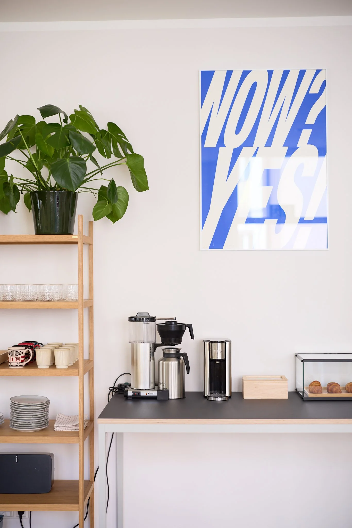 Moderner Kaffeeautomat mit Zubehör wie Tassen, Gläsern, und Broten auf einem schwarzen Arbeitstisch in einer weißen Wandküche. Ein grüner Topf mit einer großen Monstera-Pflanze steht auf einem Holzküchenregal.