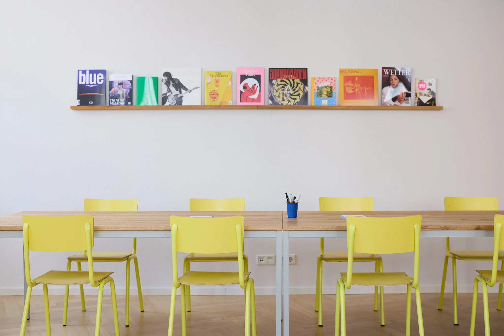 Heller Raum mit gelben Stühlen, einem langen Tisch und einer weißen Wand. Auf der Wand befindet sich eine schmale Holzablage mit mehreren bunten Magazinen, darunter auch ein Magazin mit dem Titel 'blue'.