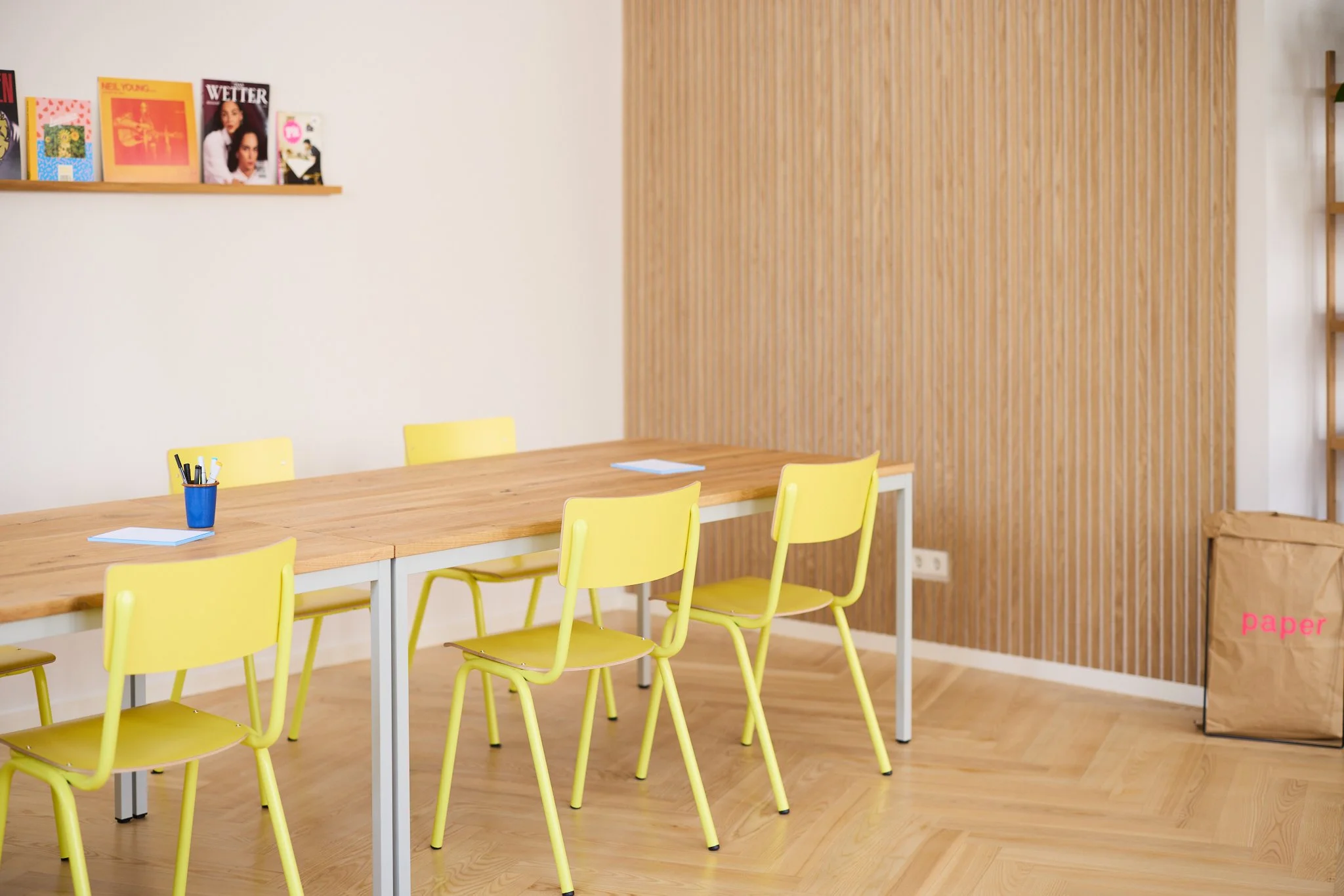 Ein heller Raum mit einer langen Holztafel, umgeben von gelben Stühlen. An der Wand hängen Magazine und Poster. Es gibt eine Holzvertäfelung an einer Wand und eine Papiertüte steht in der Ecke. Auf dem Tisch liegt ein Blatte und ein Stifthalter.
