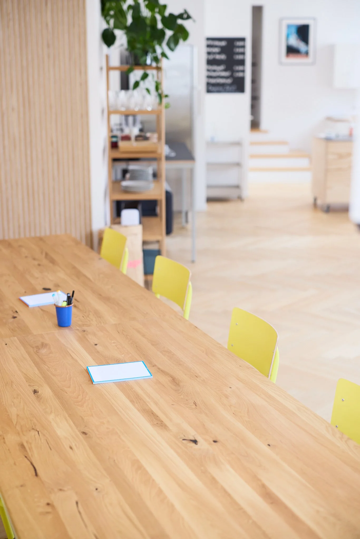 Ein heller, großer Konferenztisch aus Holz mit gelben Stühlen. Im Hintergrund eine Küche oder einen Gemeinschaftsraum mit einem Regal, Pflanzen und einem schwarzen Menübrett. Die Umgebung wirkt modern und freundlich.