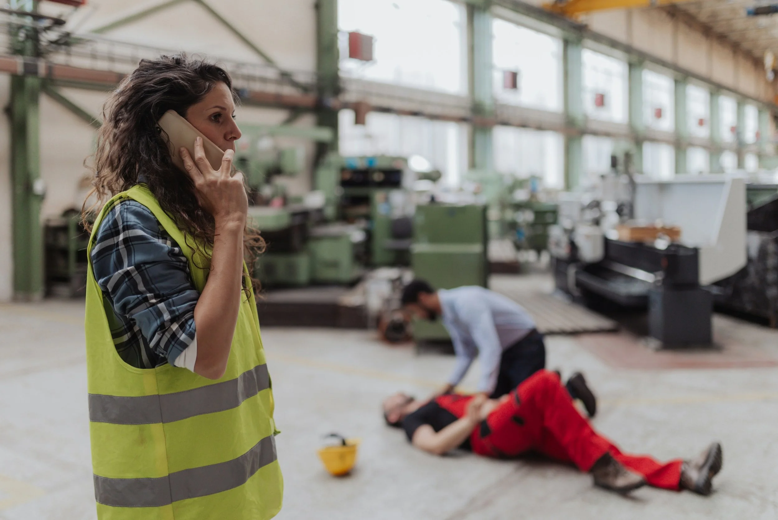 A woman in a yellow safety vest talking on a landline phone inside a large industrial or warehouse space. In the background, two people are lying on the floor, with one in a red jacket and the other in a white shirt, possibly performing a medical emergency practice or training.