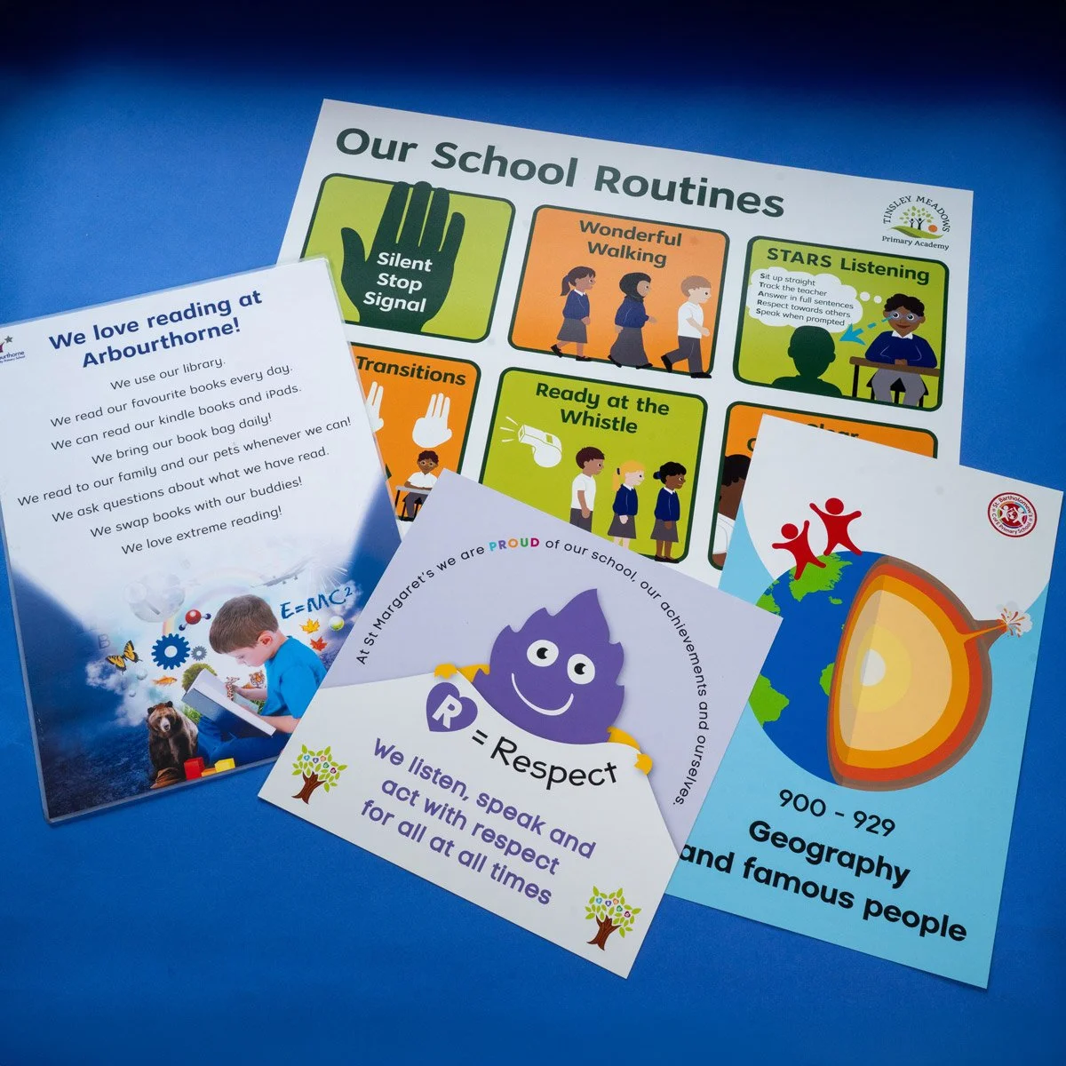 Collection of colorful school posters and flyers on a blue surface, including a school routines chart, a reading enthusiasm flyer, and a geography fact sheet.