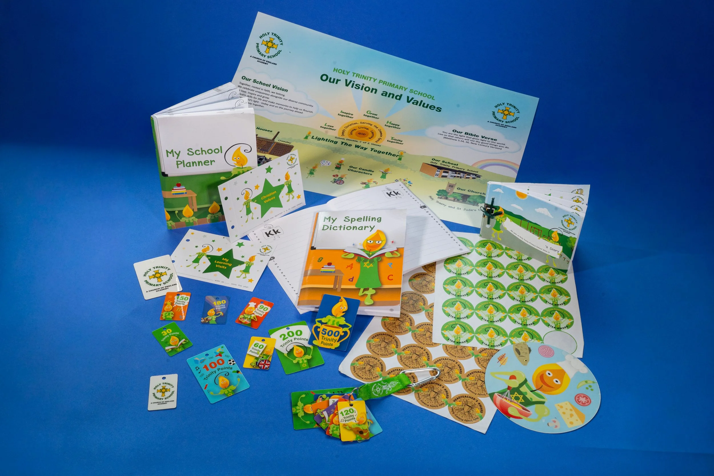 Collection of school supplies and educational materials related to Holy Trinity Primary School, including a school planner, spelling dictionary, stickers, badges, posters, and educational cards with colorful characters and points system on a blue bac