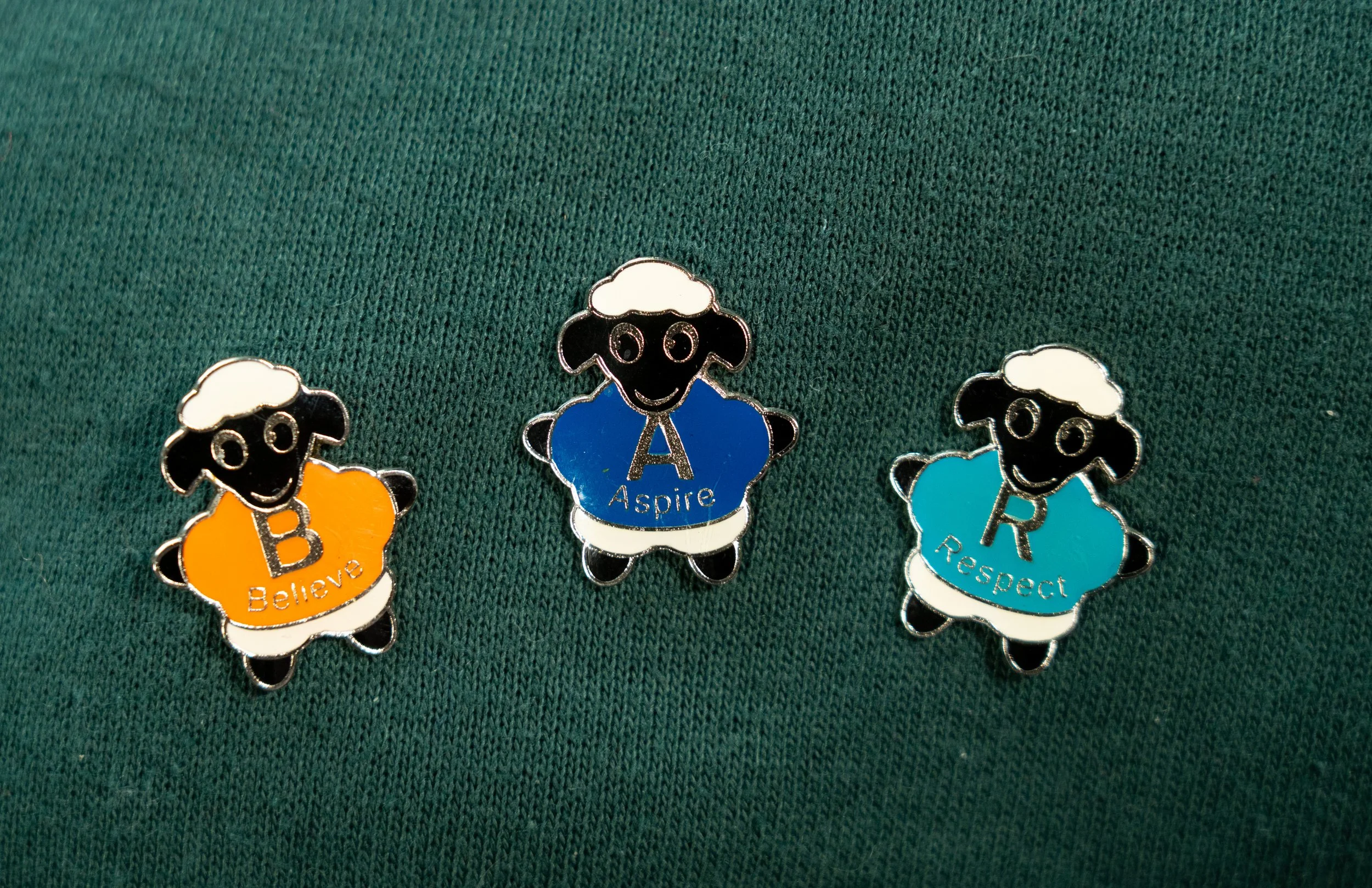 A set of enamel pins featuring cartoon sheep with white wool, black faces, and smiling faces. Each sheep wears a colored shirt with a word: 'Believe' on an orange shirt, 'Aspire' on a blue shirt, and 'Respect' on a light blue shirt.