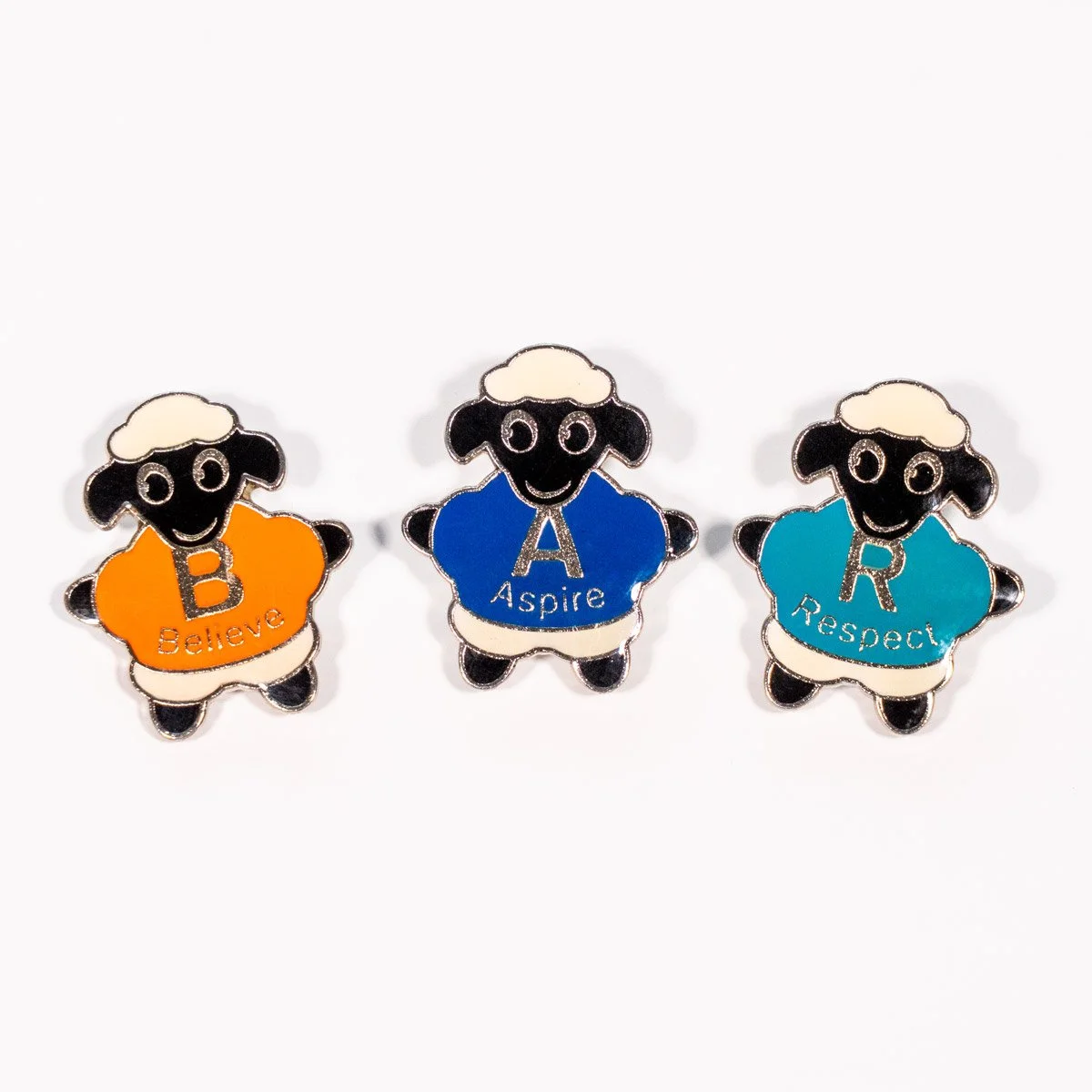Three sheep-shaped pins with words: Believe in orange, Aspire in blue, Respect in teal, each with a black face, white wool, and black legs and ears.