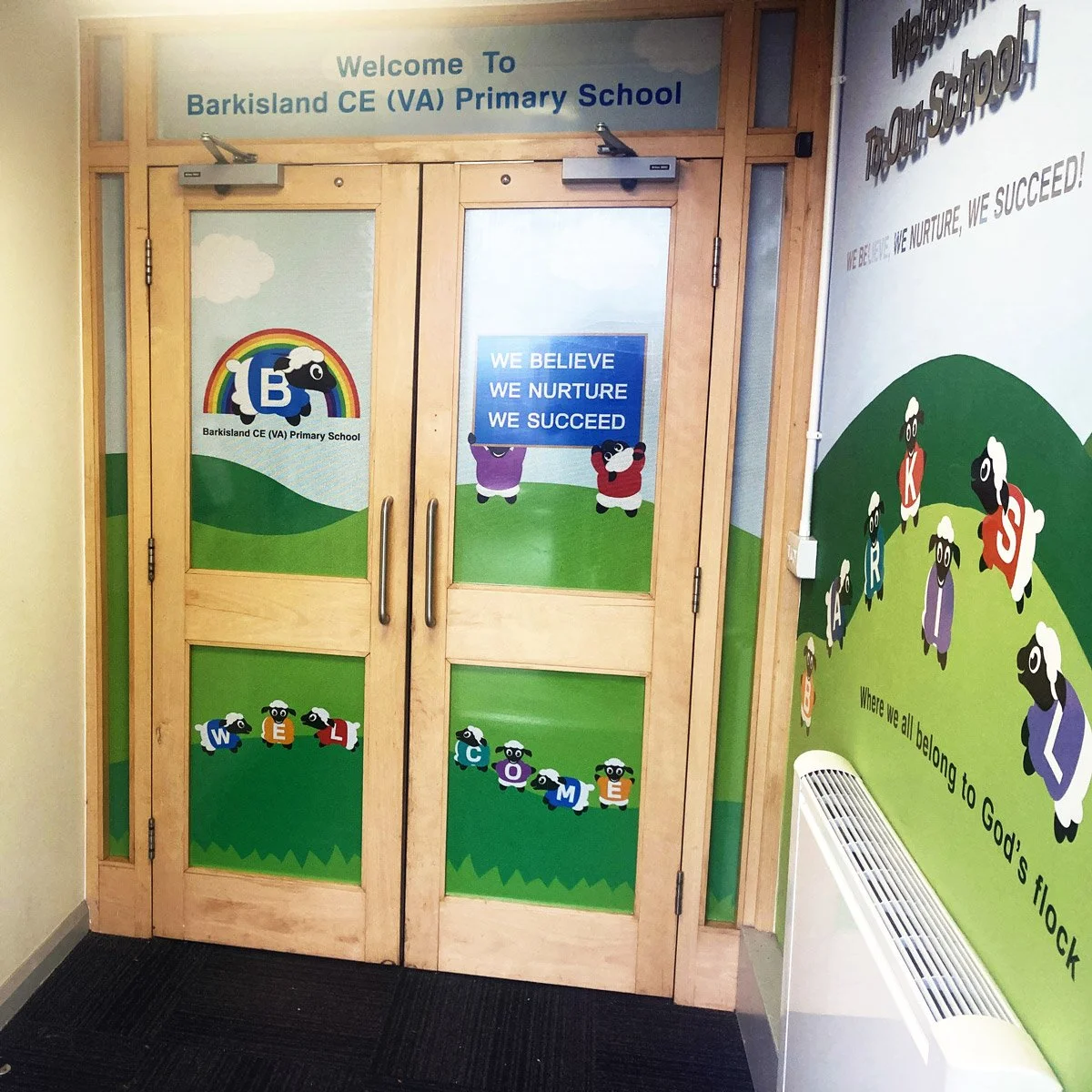 Wooden double doors at Barkisland CE (VA) Primary School with colorful cartoon sheep, each with a letter spelling 'WELCOME' on the bottom panels. The top panel above the doors has the school name and welcoming message, and another sign on the glass says, 'WE BELIEVE WE NURTURE WE SUCCEED.' The wall on the right has a mural of sheep with the message, 'Where we all belong to God's flock.'