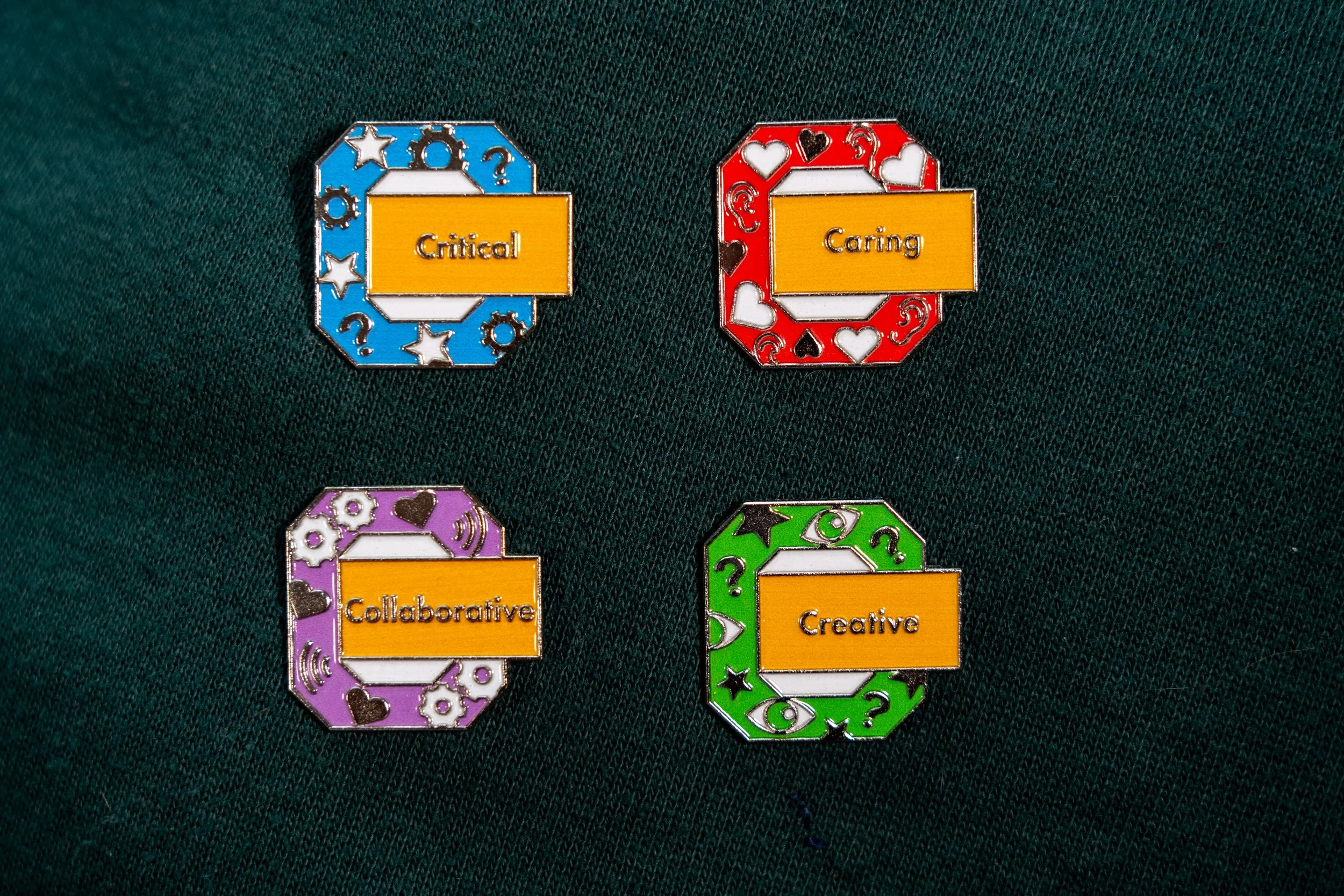 Colorful enamel pins with words 'Critical,' 'Caring,' 'Collaborative,' and 'Creative' on yellow backgrounds, attached to dark fabric.