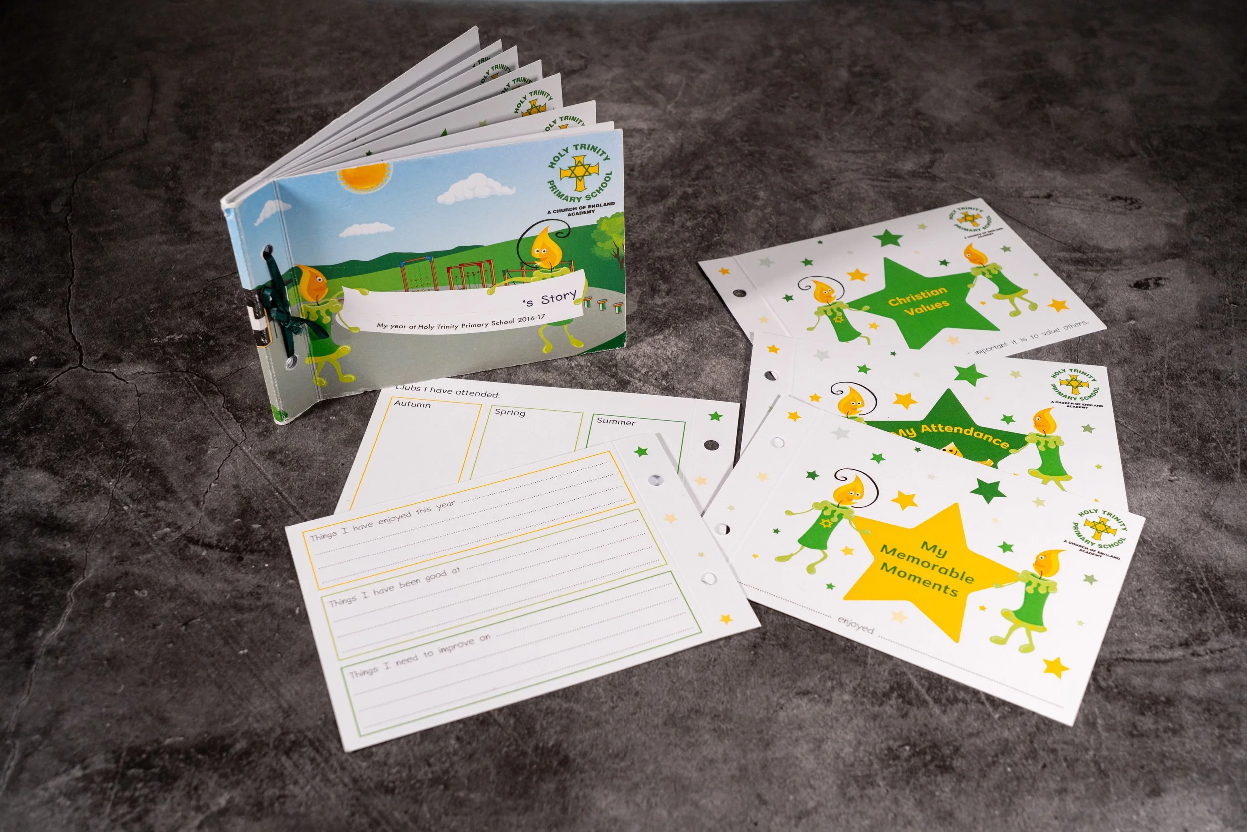 A small booklet with a blue cover featuring cartoon characters and the logo of Holy Trinity Primary School, with accompanying sheets of paper about Christian values and memorable moments, all placed on a dark surface.