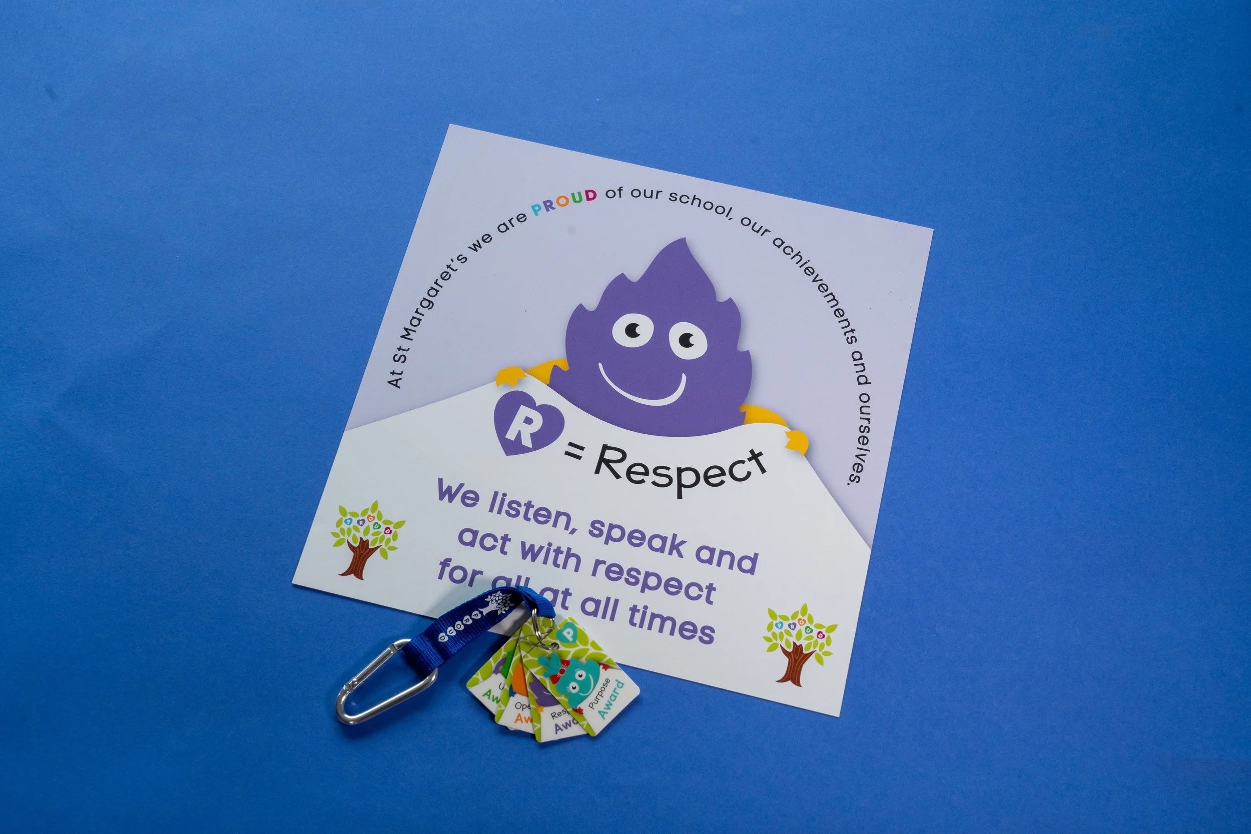 A colorful school badge with a cartoon purple flame character holding a purple heart labeled "Respect". The badge has a rainbow-colored word "PROUD" and text about respecting and acting with respect. There are also small tags with the words "Awesome"