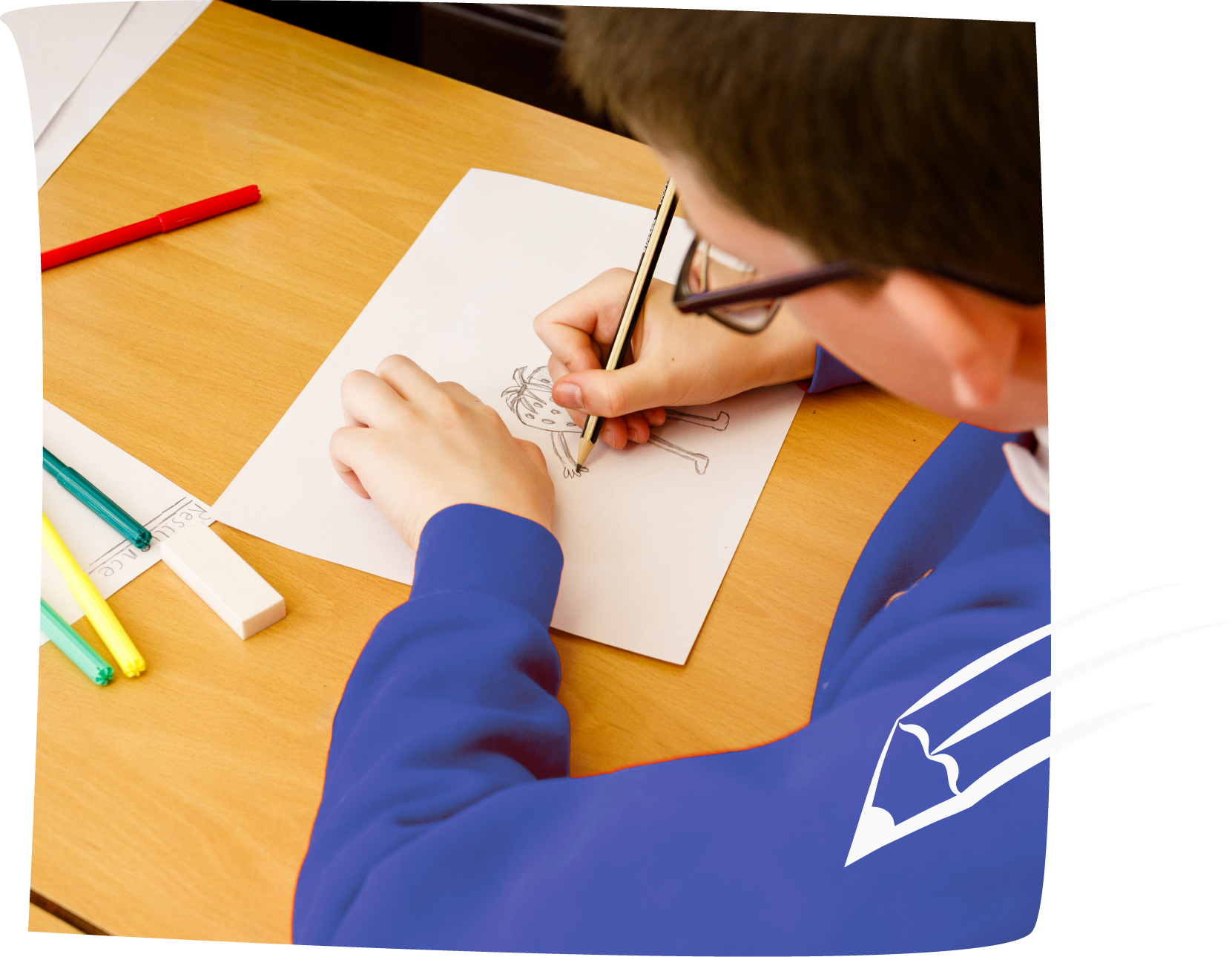 A school boy  in a blue school sweatshirt is drawing a character design with a pencil. There are felt tips and an eraser on the desk nearby. The edge of another pupil's drawing can be glimpsed opposite