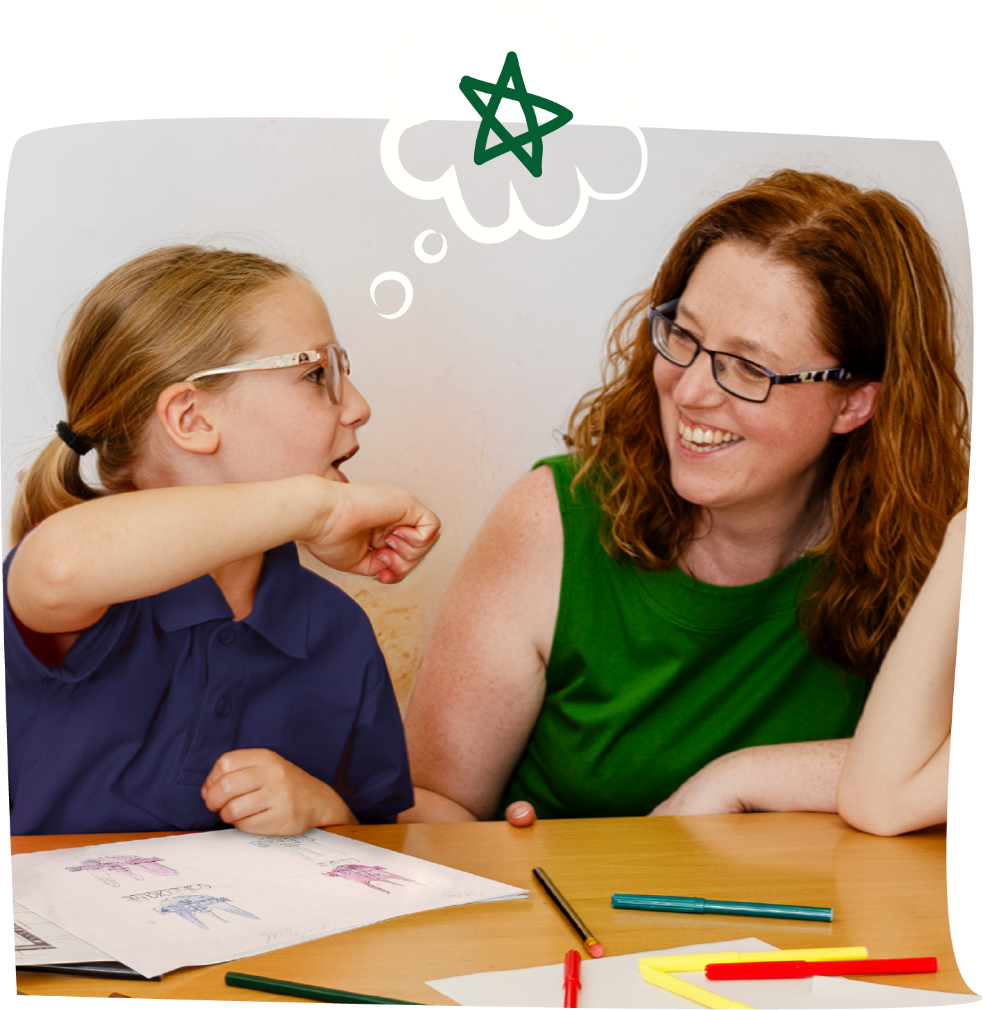 A young girl and an adult woman sit at a table, having a conversation. The girl is wearing glasses and a navy blue shirt, and the woman is wearing glasses and a green sleeveless top, both smiling. On the table, there are drawings, colored markers, and a pen.