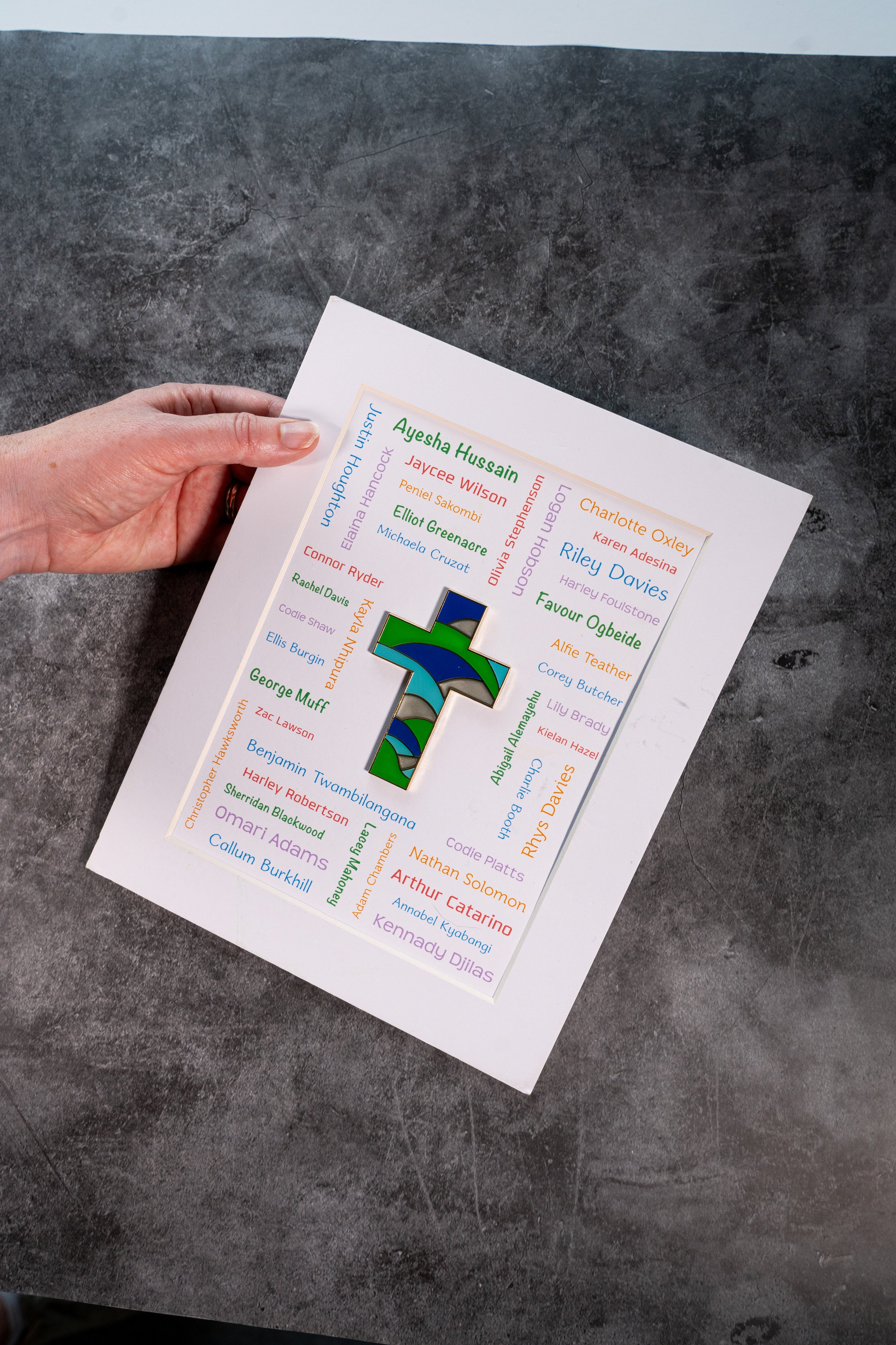A person holding a printed sheet with a list of names in different colors, centered around a decorated cross in the middle, on a dark gray surface.