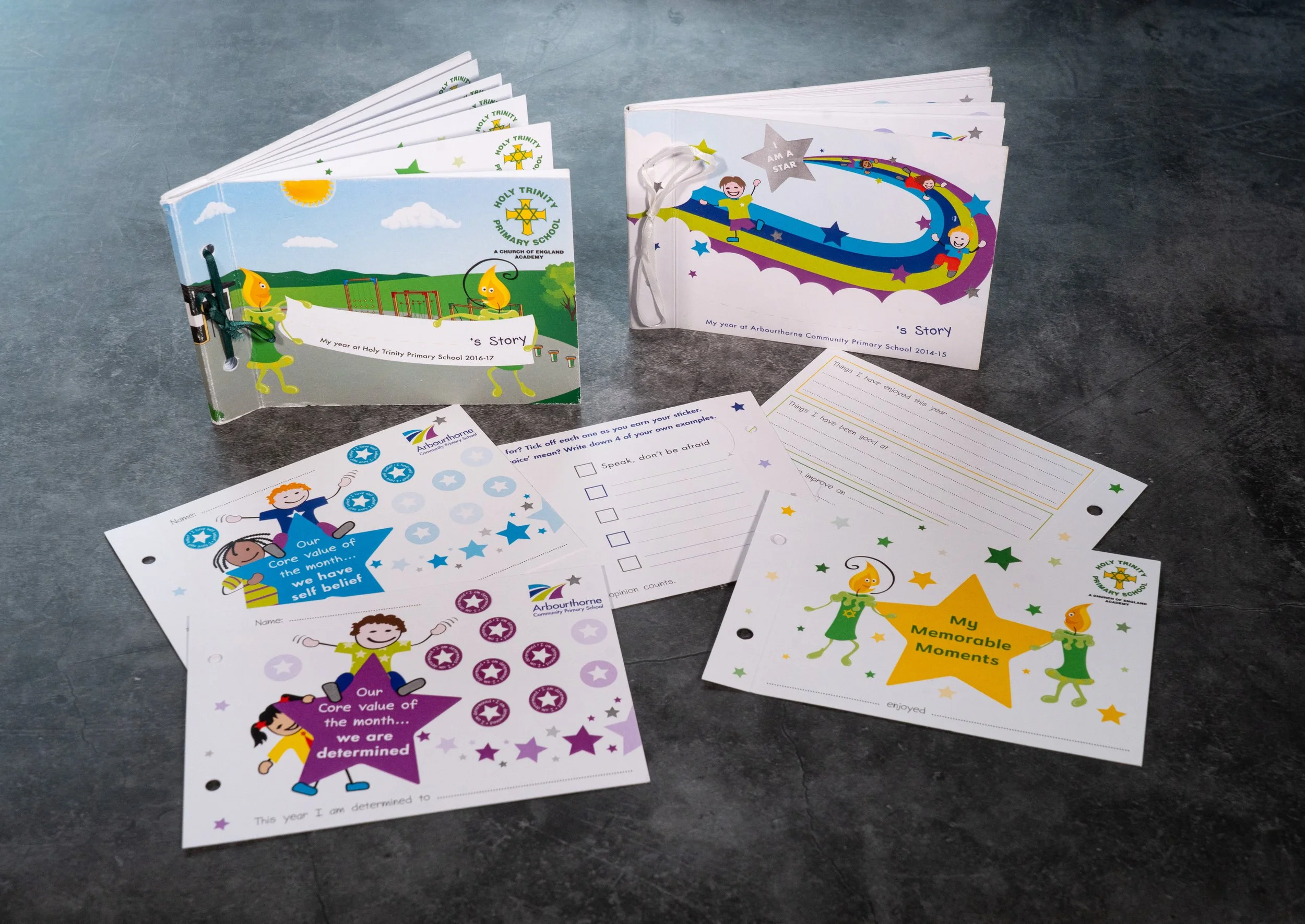 Two colorful school storybooks with cartoon characters on the covers are displayed on a gray surface. One book has a green and blue cover featuring two animated characters and the Holy Trinity Primary School logo, while the other has a white cover wi