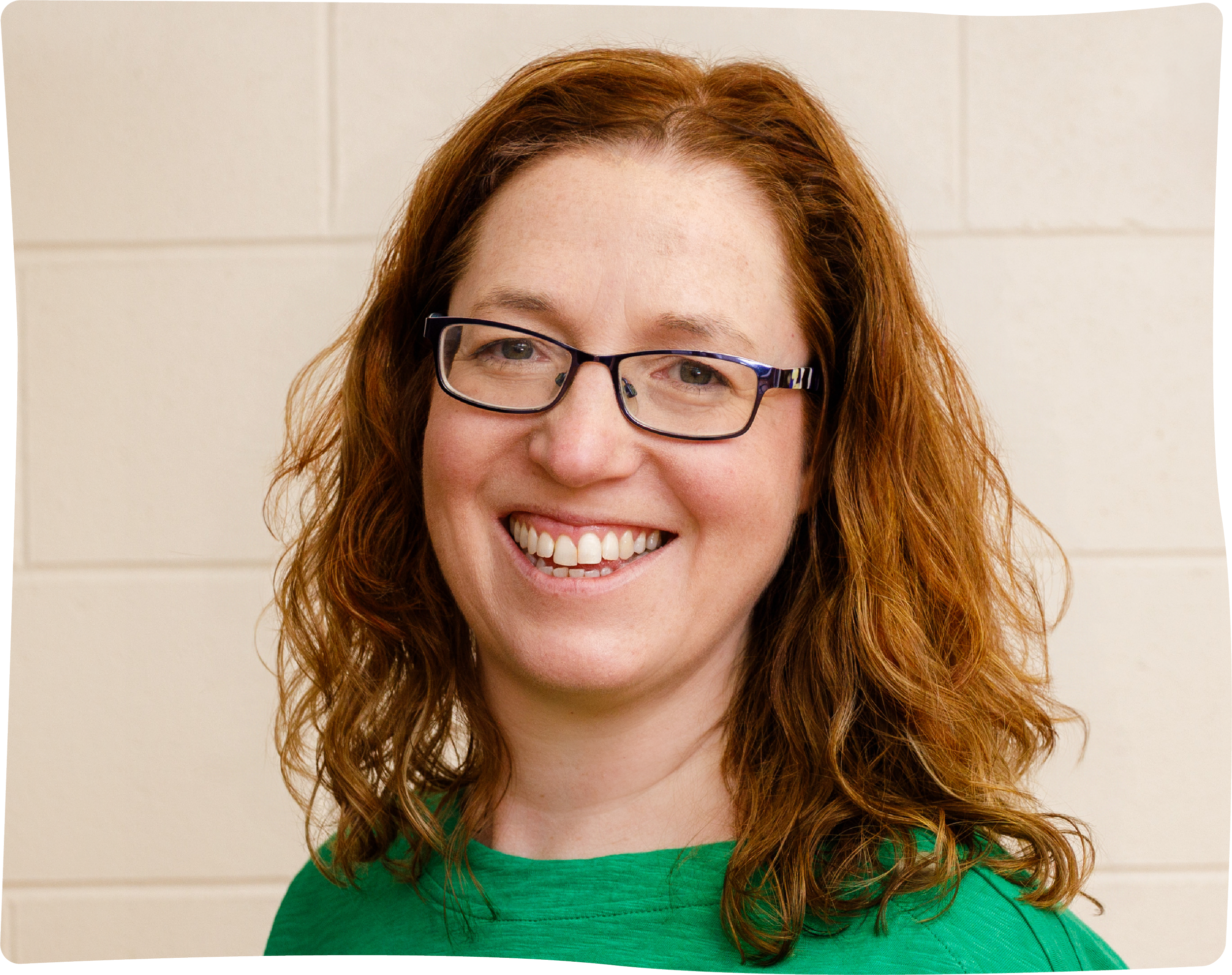 Close-up of a woman with red, curly hair wearing glasses and a green shirt, smiling against a cream background.